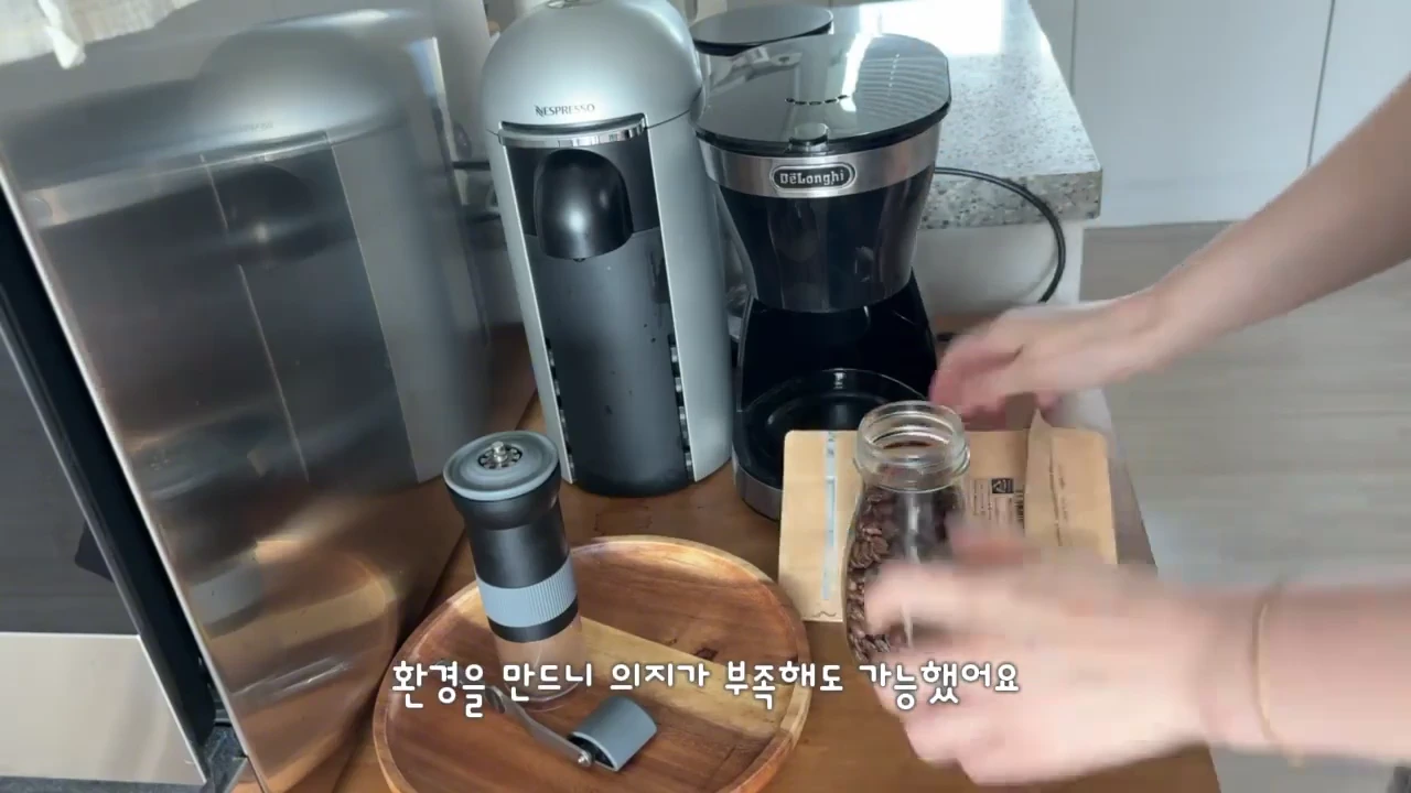 주방 조리대 위에 커피 머신 두 대와 원두가 담긴 유리병이 놓여 있고, 사람의 손이 유리병을 만지고 있는 모습