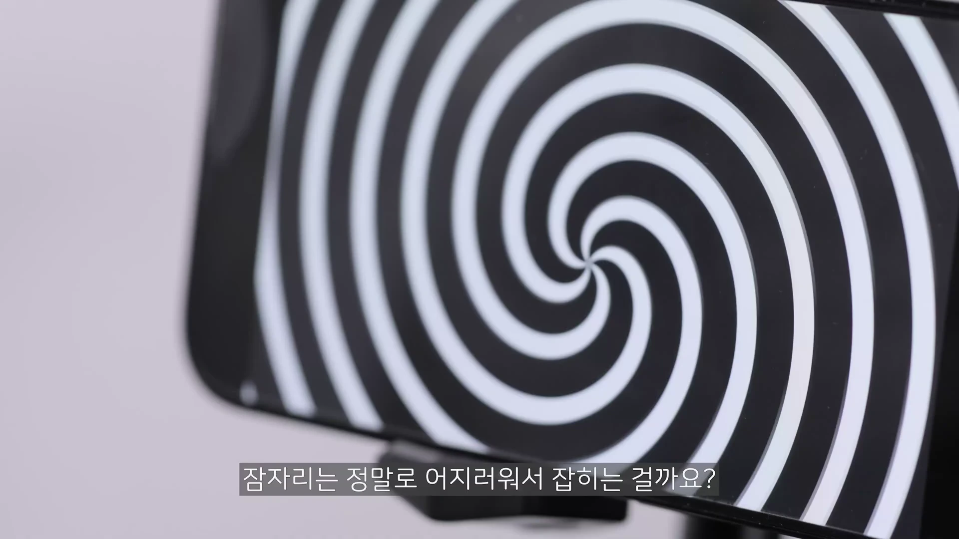 스마트폰 화면에 흑백 소용돌이 무늬가 회전하는 영상이 재생되고 있으며, 하단에는 한국어 자막이 표시되어 있습니다.