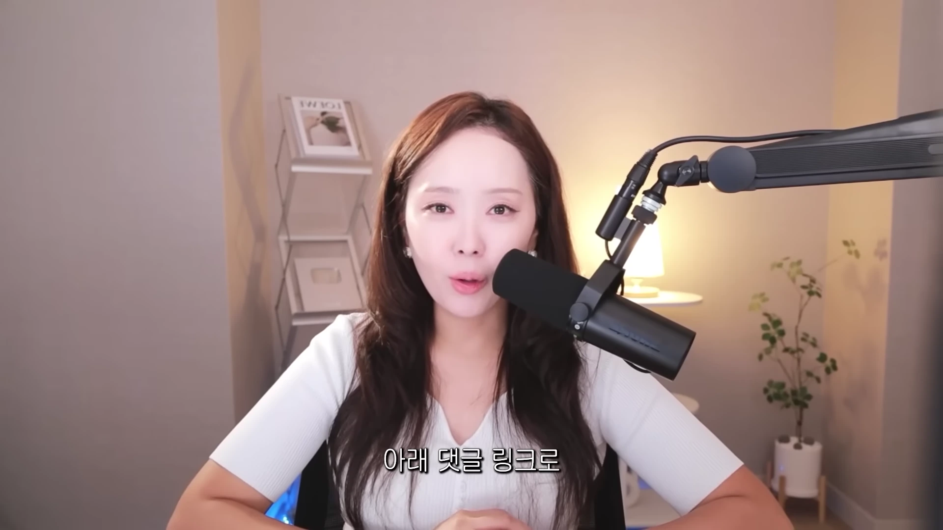 흰색 상의를 입은 여성이 책상 앞에 앉아 검은색 마이크 앞에 대고 이야기하고 있으며, 배경에는 조명과 선반 등 아늑한 실내 공간이 보인다.