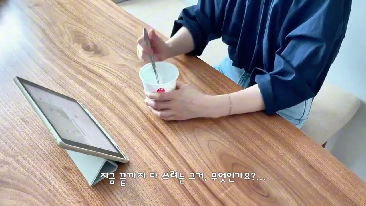 나무 식탁 앞에 앉아 태블릿을 보며 숟가락으로 용기 안의 내용물을 젓고 있는 사람의 손.