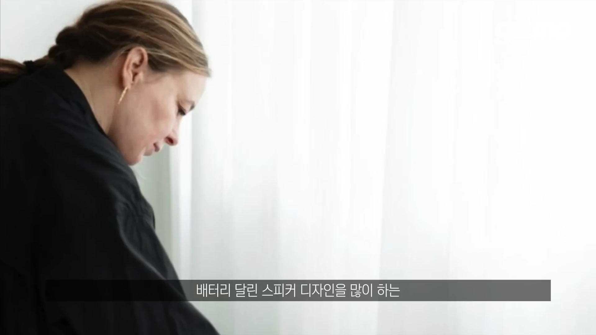 검은색 상의를 입은 여성이 흰색 커튼 앞에 서 있는 옆모습이 담긴 영상 캡처 화면이며 하단에 '배터리 달린 스피커 디자인을 많이 하는'이라는 자막이 표시되어 있습니다.