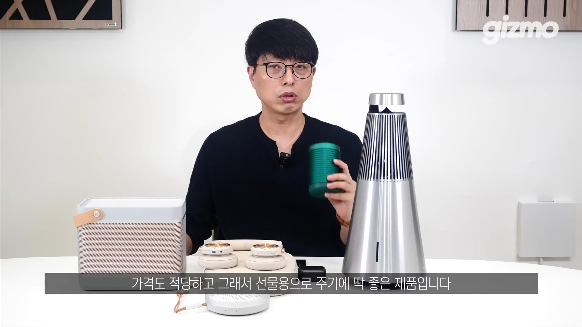 안경을 쓴 남성이 스튜디오 테이블 앞에 앉아 손에 작은 녹색 원통형 스피커를 들고 설명하고 있으며, 테이블 위에는 다양한 모양과 색상의 뱅앤올룹슨 스피커와 헤드폰이 놓여 있습니다.