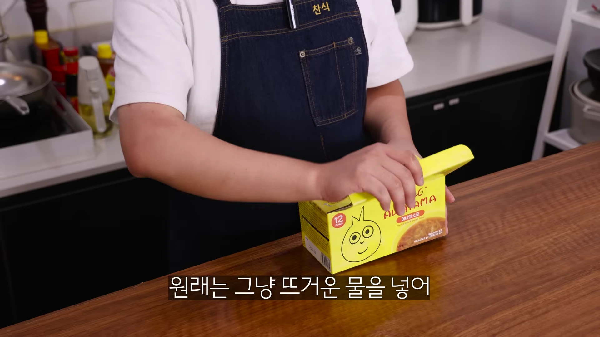 앞치마를 두른 요리사가 양파 캐릭터가 그려진 노란색 동결건조 양파 스프 상자를 양손으로 잡고 있는 모습