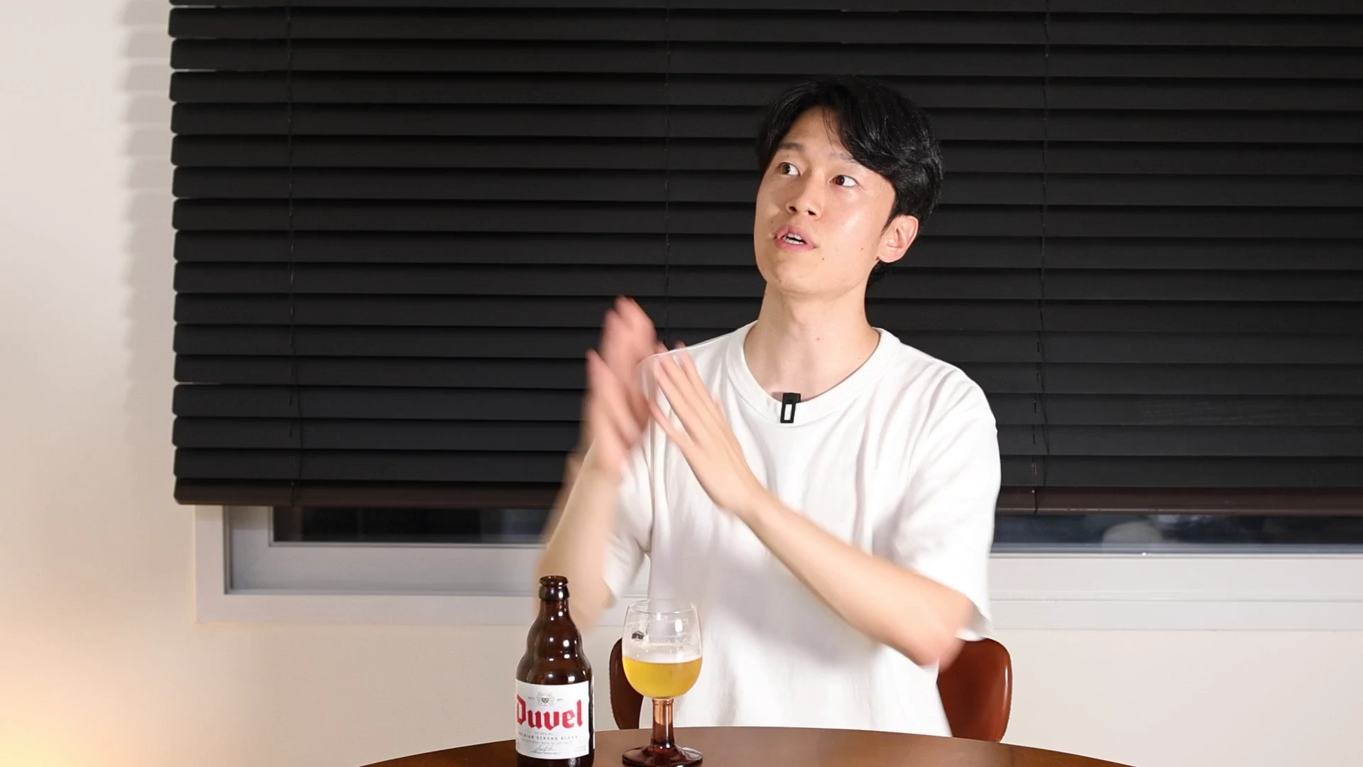 흰색 티셔츠를 입은 남성이 검은색 블라인드 배경 앞에서 손짓하며 이야기를 하고 있고, 테이블 위에는 맥주병과 잔이 놓여 있습니다.