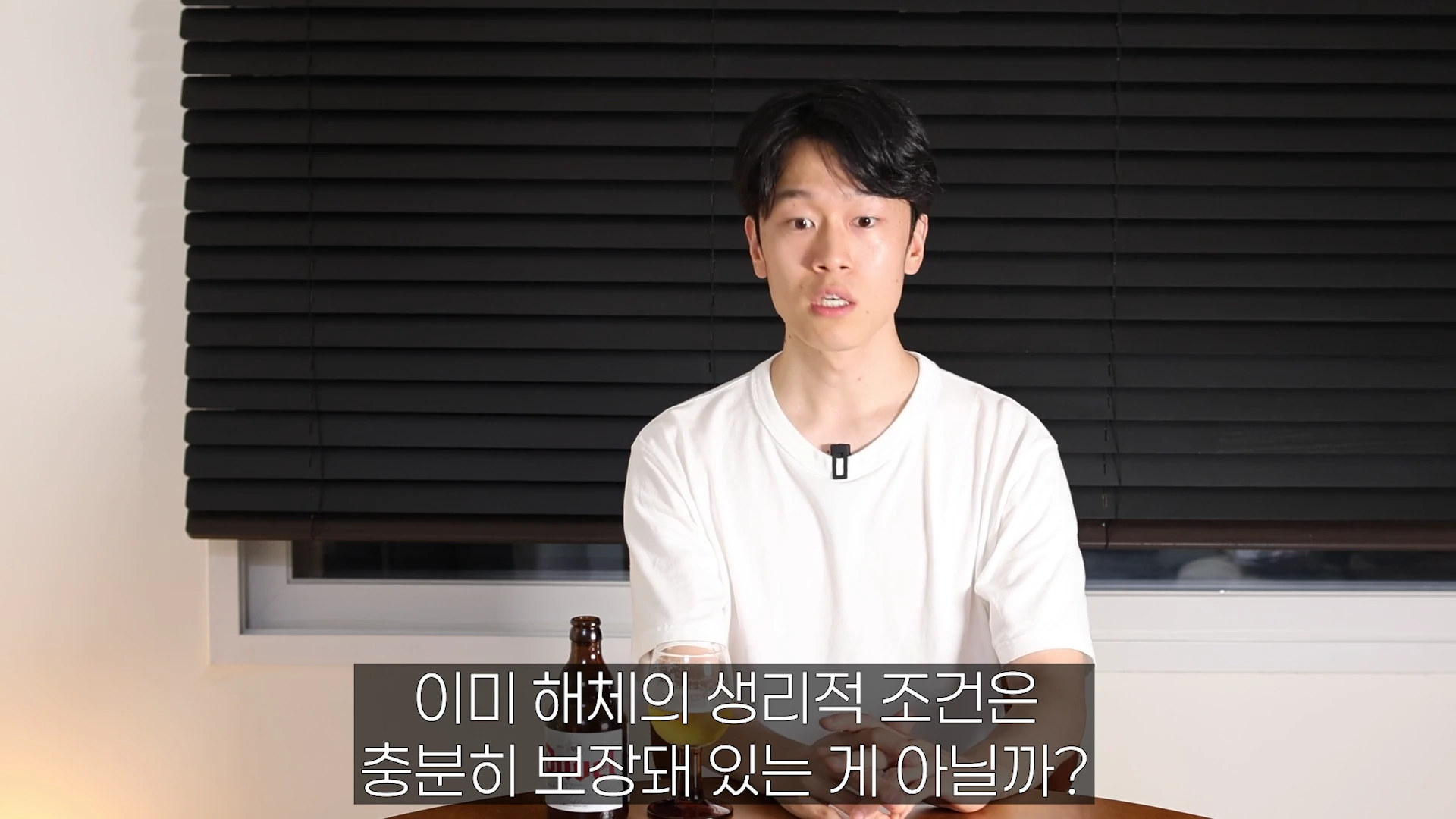 검은색 블라인드 배경 앞에 흰색 반소매 티셔츠를 입은 젊은 남성이 앉아 있고, 앞에는 맥주병과 잔이 놓여 있으며 화면 하단에는 자막이 떠 있습니다.