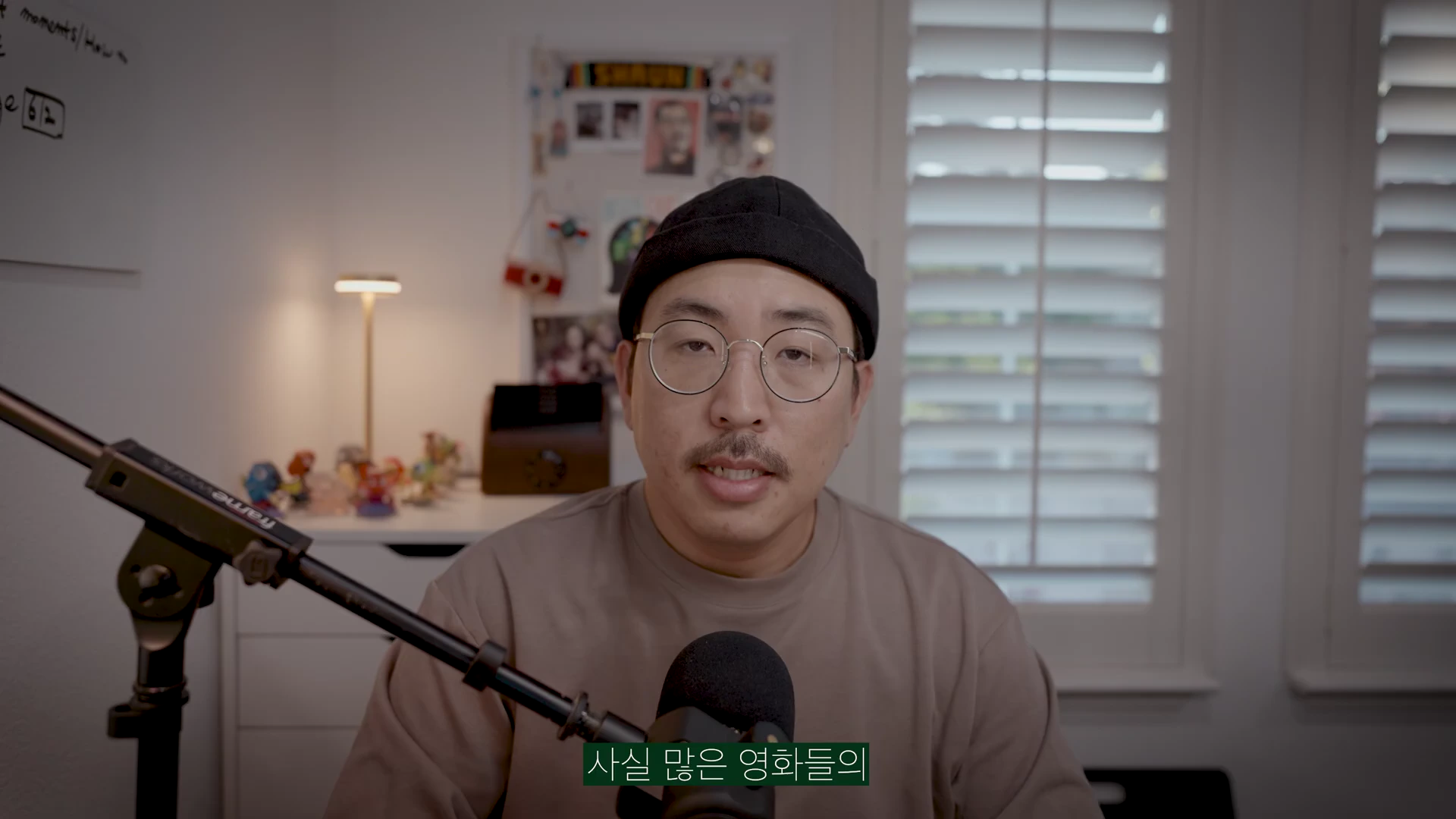 실내에서 검은색 비니와 안경을 쓴 남성이 마이크 앞에 앉아 정면을 보며 이야기를 하고 있는 모습