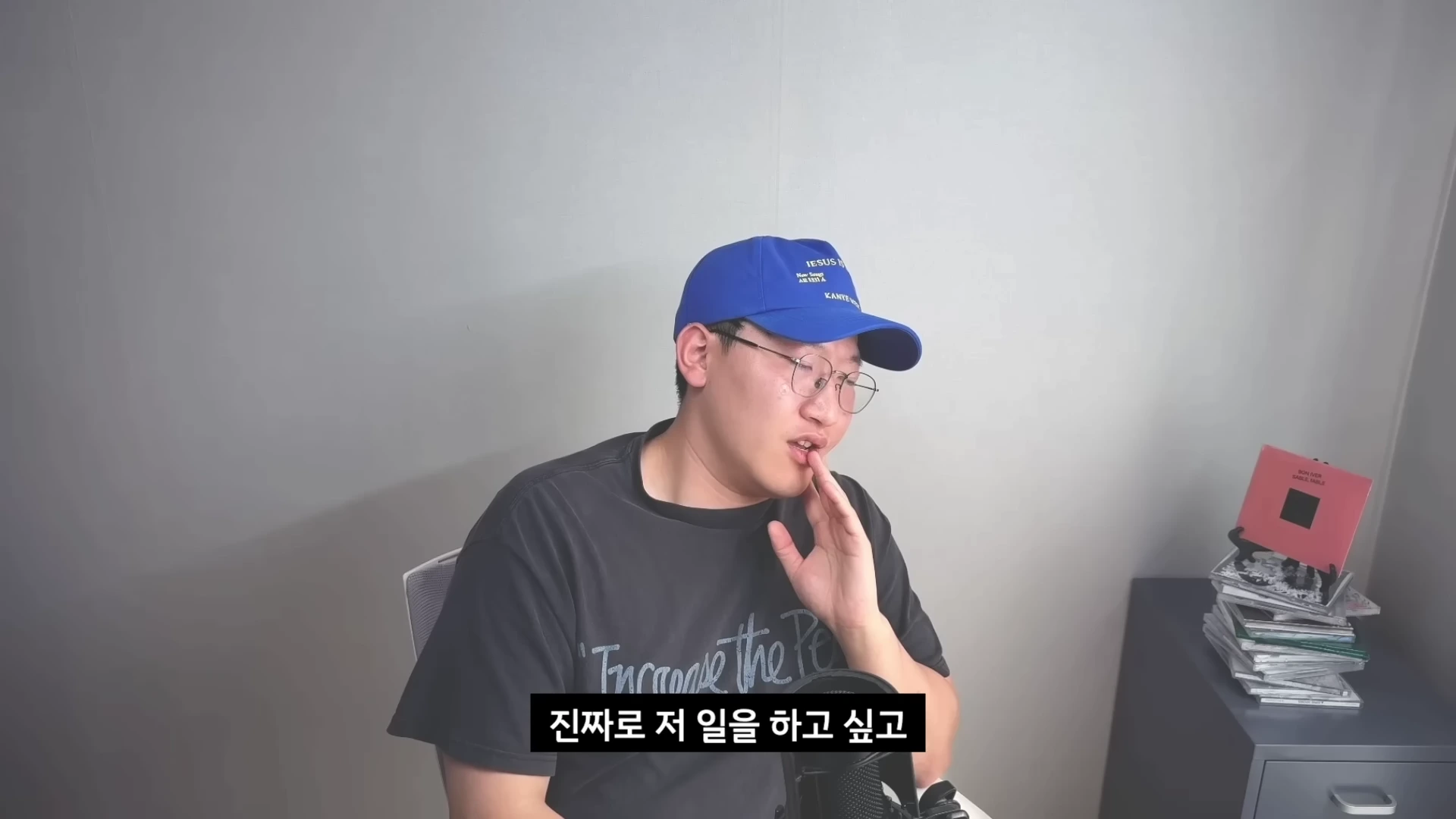 파란색 야구 모자와 안경을 쓴 남성이 마이크 앞에서 고민하는 듯한 표정으로 손을 턱에 대고 이야기를 하고 있는 모습