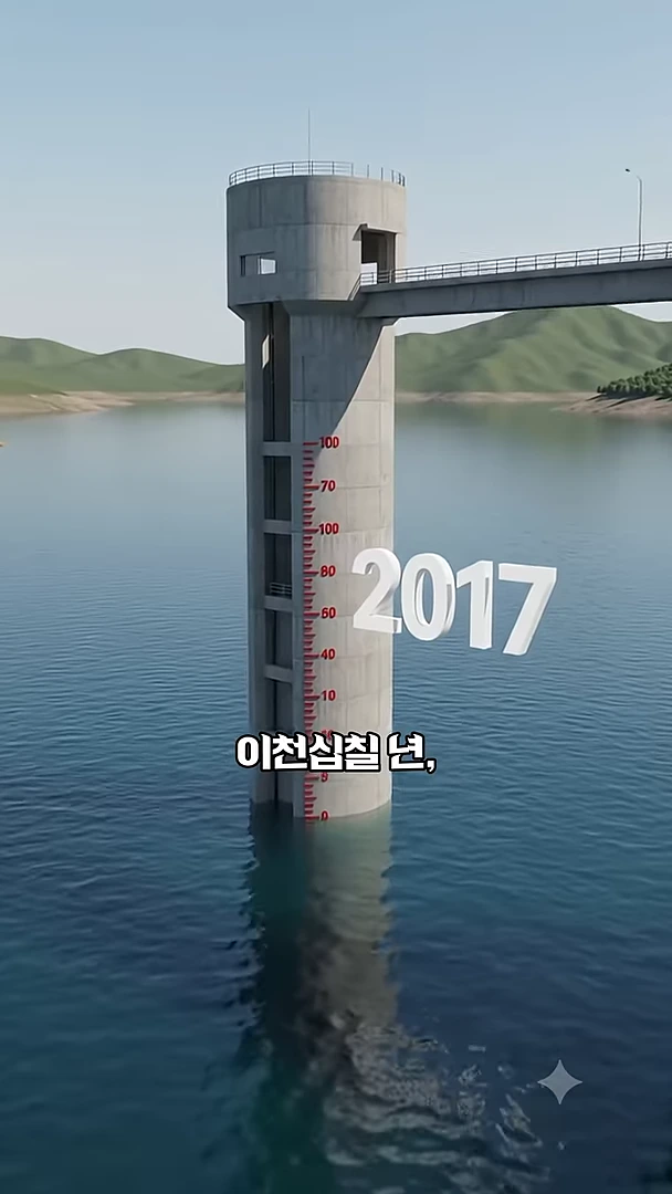 호수 중앙에 세워진 원통형의 회색 선택취수탑이 보이며 탑의 표면에는 수심을 나타내는 숫자와 눈금이 그려져 있고 옆으로 연결된 다리가 주변 산과 함께 보입니다.
