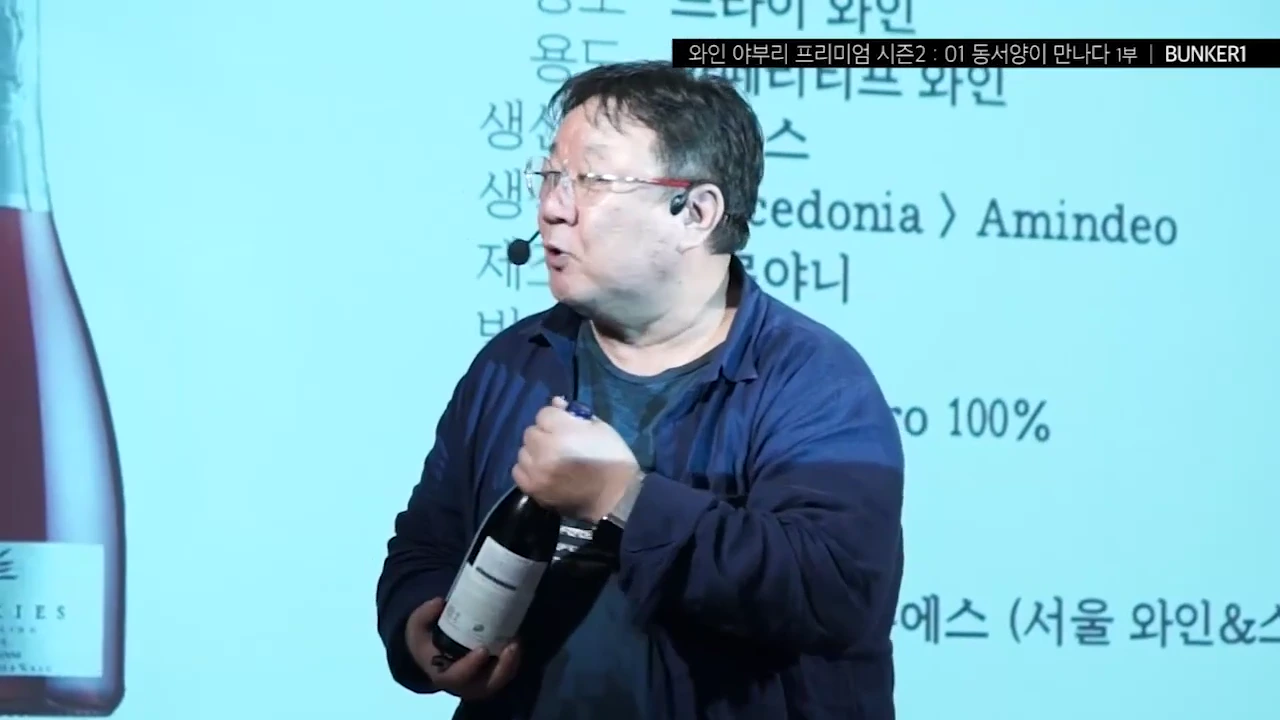 안경을 쓴 중년 남성이 강의실에서 한 손으로 와인 병을 들고 포일 캡 부분을 돌려보는 시연을 하고 있다.
