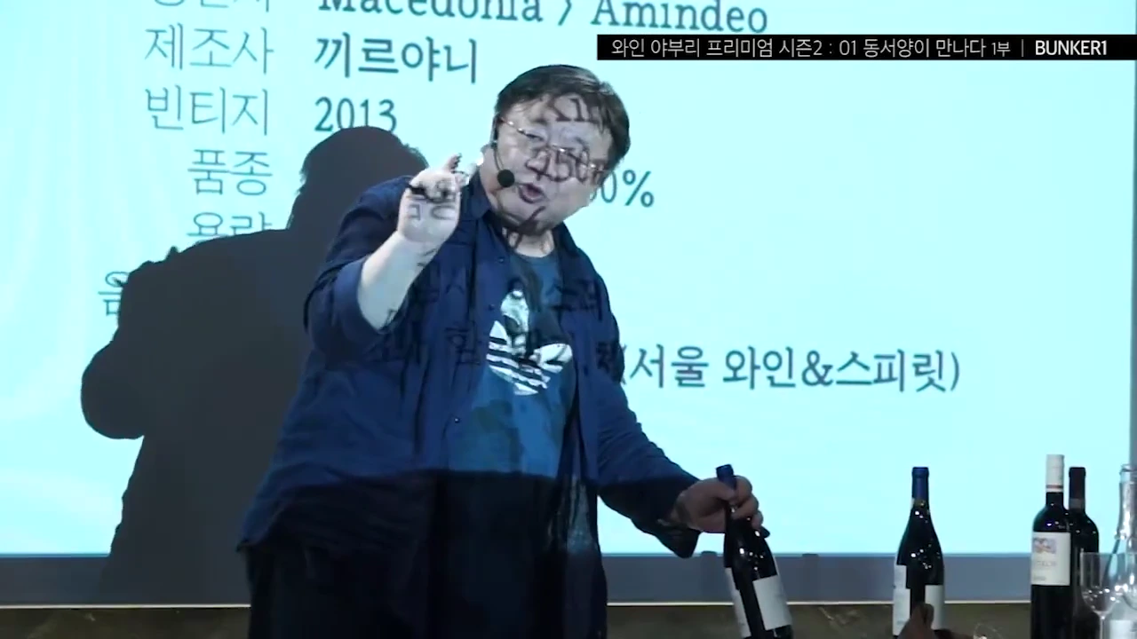 안경을 쓴 남성이 무대 위에서 소믈리에 나이프를 손에 들고 와인병을 활용해 코르크를 뽑는 시연을 하고 있으며, 뒤로는 와인 정보가 적힌 스크린이 보입니다.