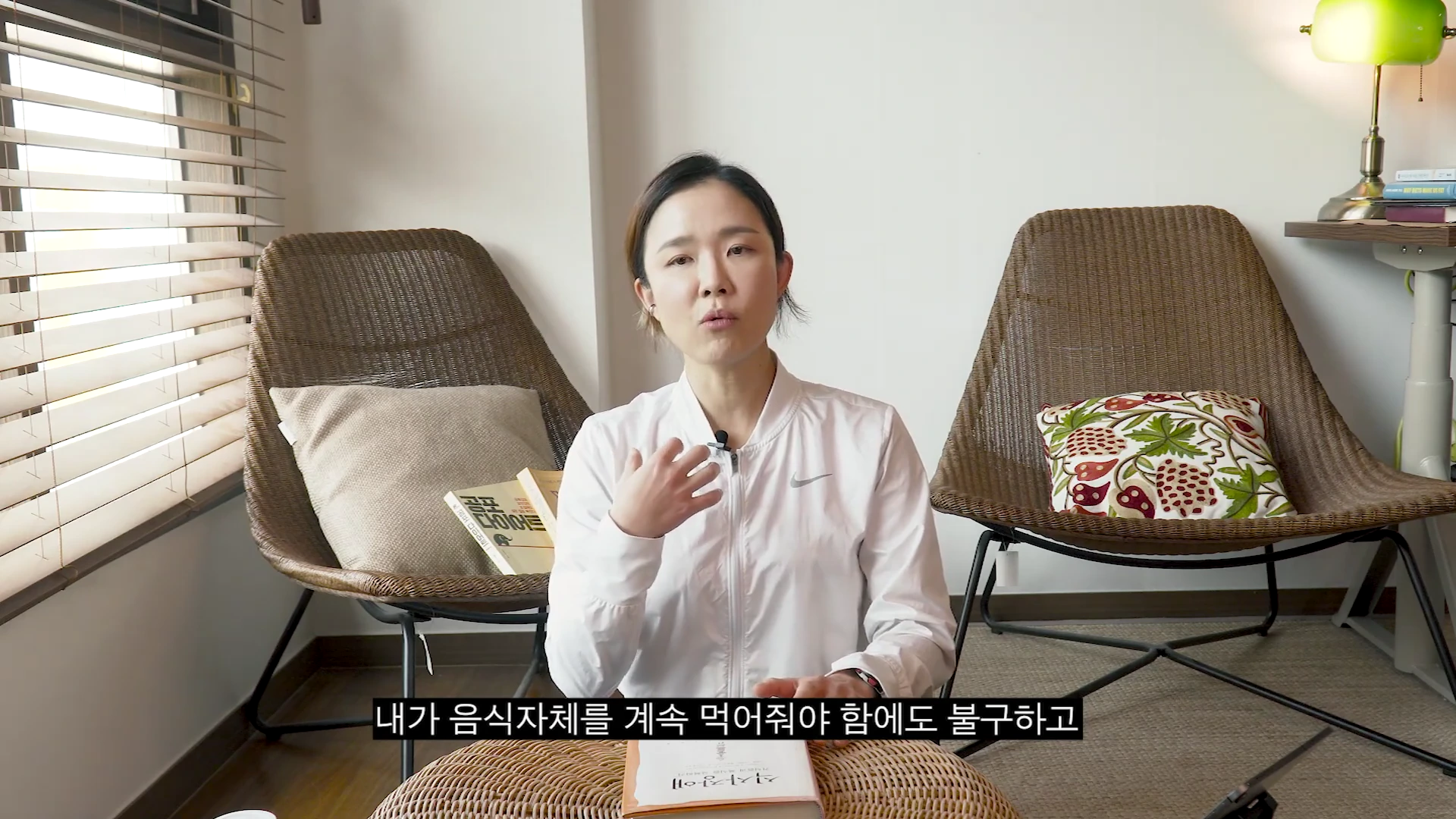 흰색 집업 재킷을 입은 여성이 의자에 앉아 손짓을 하며 무언가를 설명하고 있고 앞에는 책이 놓여 있는 스튜디오 풍경.