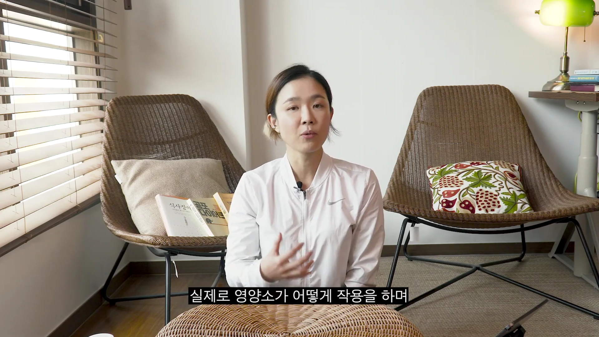 실내에서 흰색 집업 재킷을 입은 여성이 의자에 앉아 손짓하며 이야기하고 있는 모습.