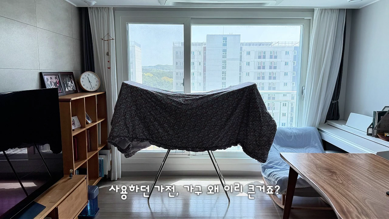 넓은 창 앞에 빨래 건조대가 놓여 있고, 왼쪽에는 거실장과 책장이, 오른쪽에는 나무 식탁과 의자가 배치된 아파트 거실 풍경.