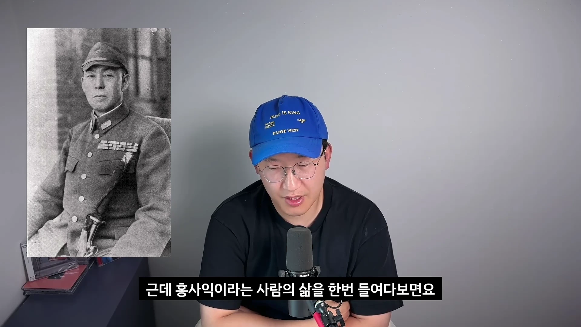 파란 모자를 쓴 남성이 마이크 앞에서 이야기를 하고 있고, 화면 왼쪽에는 군복을 입은 남성의 흑백 사진이 배치되어 있다.