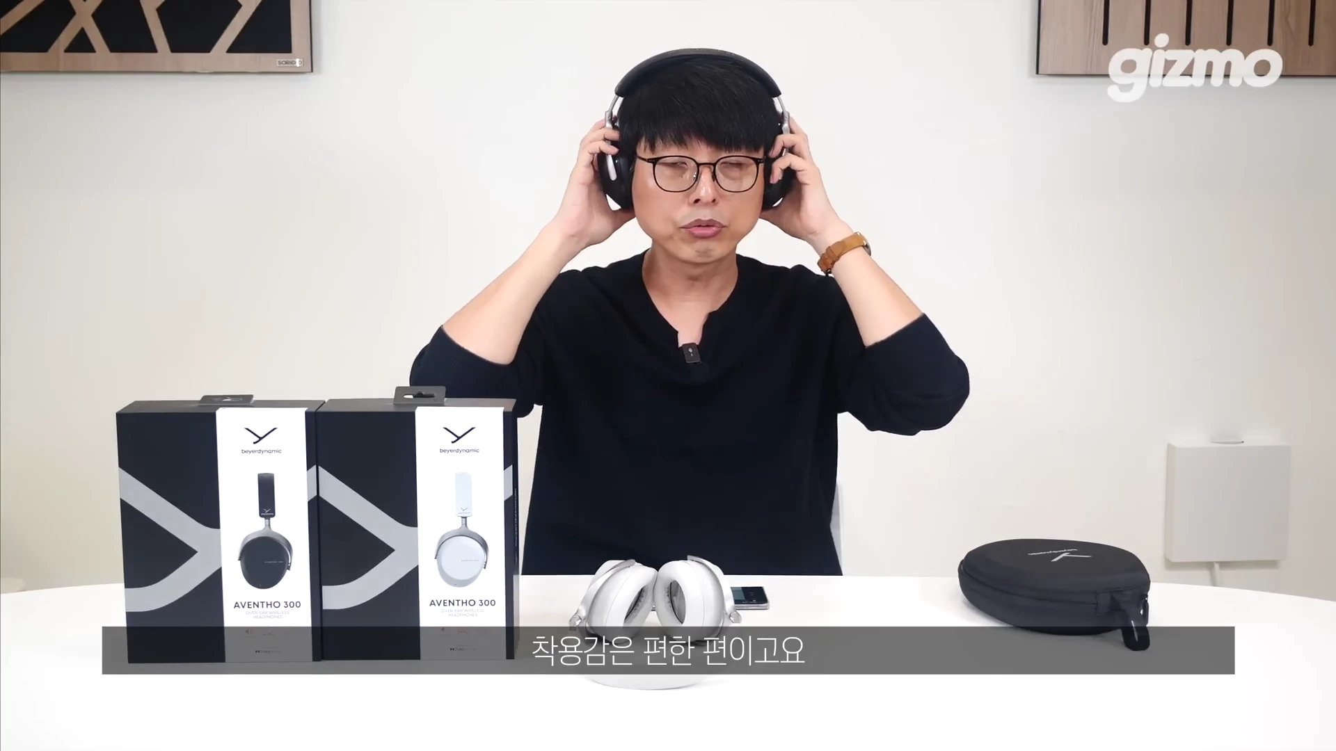안경을 쓴 남성이 헤드폰을 착용해 보며 제품의 착용감을 확인하고 있고, 테이블 위에는 제품 패키지 상자와 파우치가 놓여 있다.