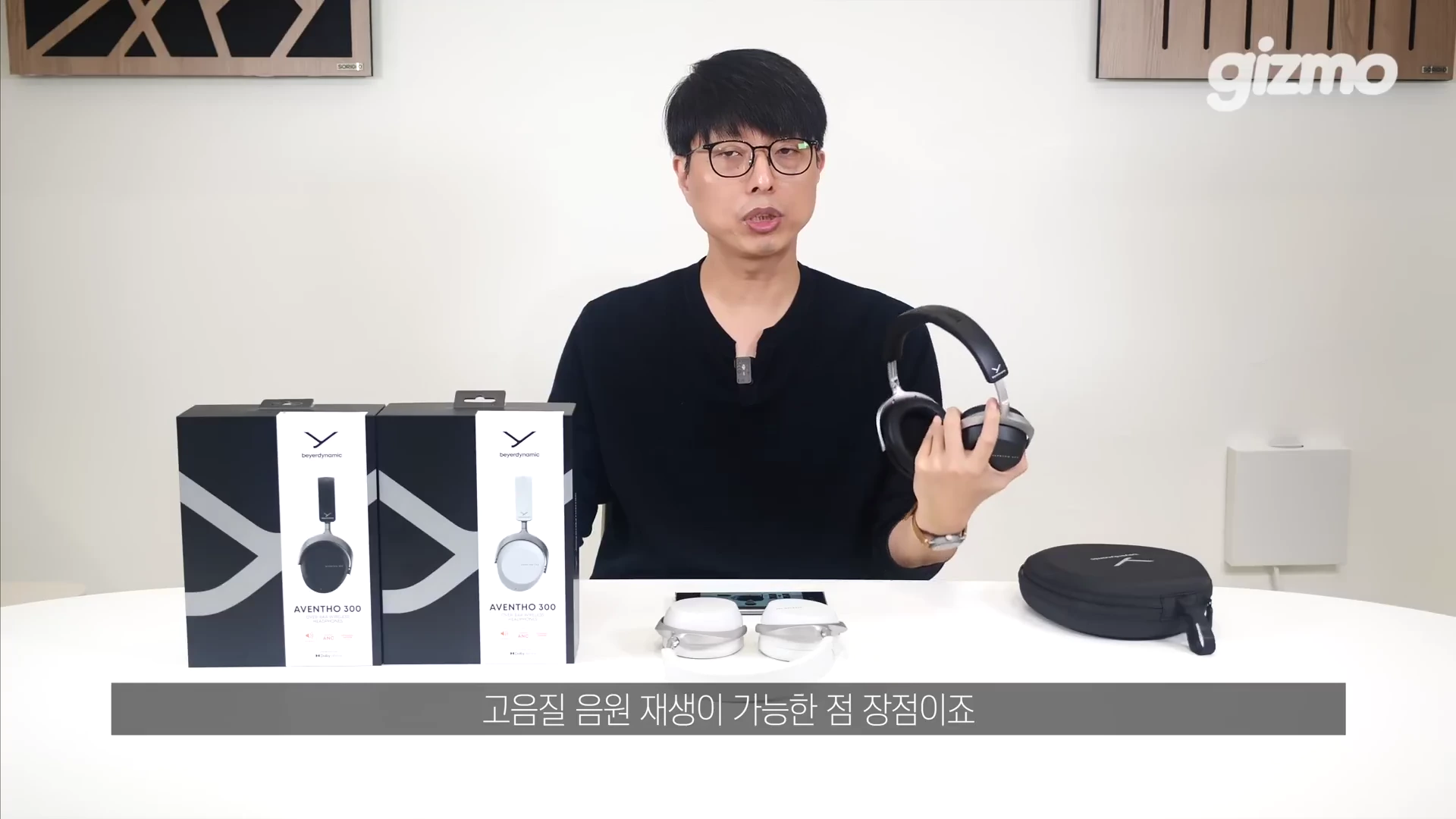 안경을 쓴 남성이 스튜디오 테이블 앞에 앉아 무선 헤드폰을 손에 들고 제품에 대해 설명하고 있다. 테이블 위에는 헤드폰 제품 상자 두 개와 헤드폰 케이스가 놓여 있다.