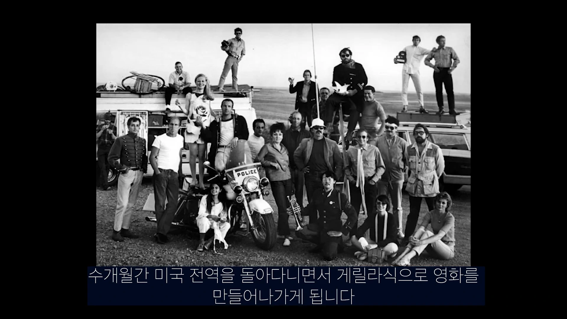 흑백 사진 속에 다양한 복장을 한 수십 명의 영화 제작진이 야외에 주차된 차량과 오토바이를 배경으로 나란히 서 있거나 앉아 있는 모습.