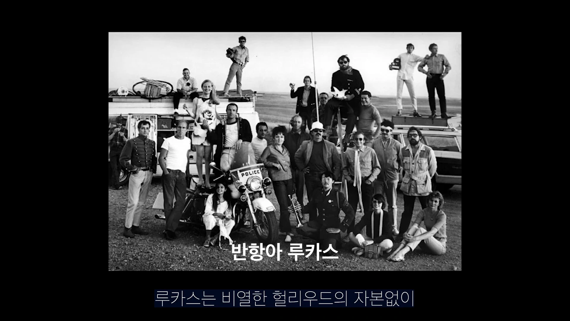 흑백 사진 속에 조지 루카스를 포함한 다수의 영화 제작진과 배우들이 경찰 오토바이와 차량 주변에 서 있거나 앉아 있는 모습. 하단에 '반항아 루카스'라는 한국어 자막이 적혀 있음.