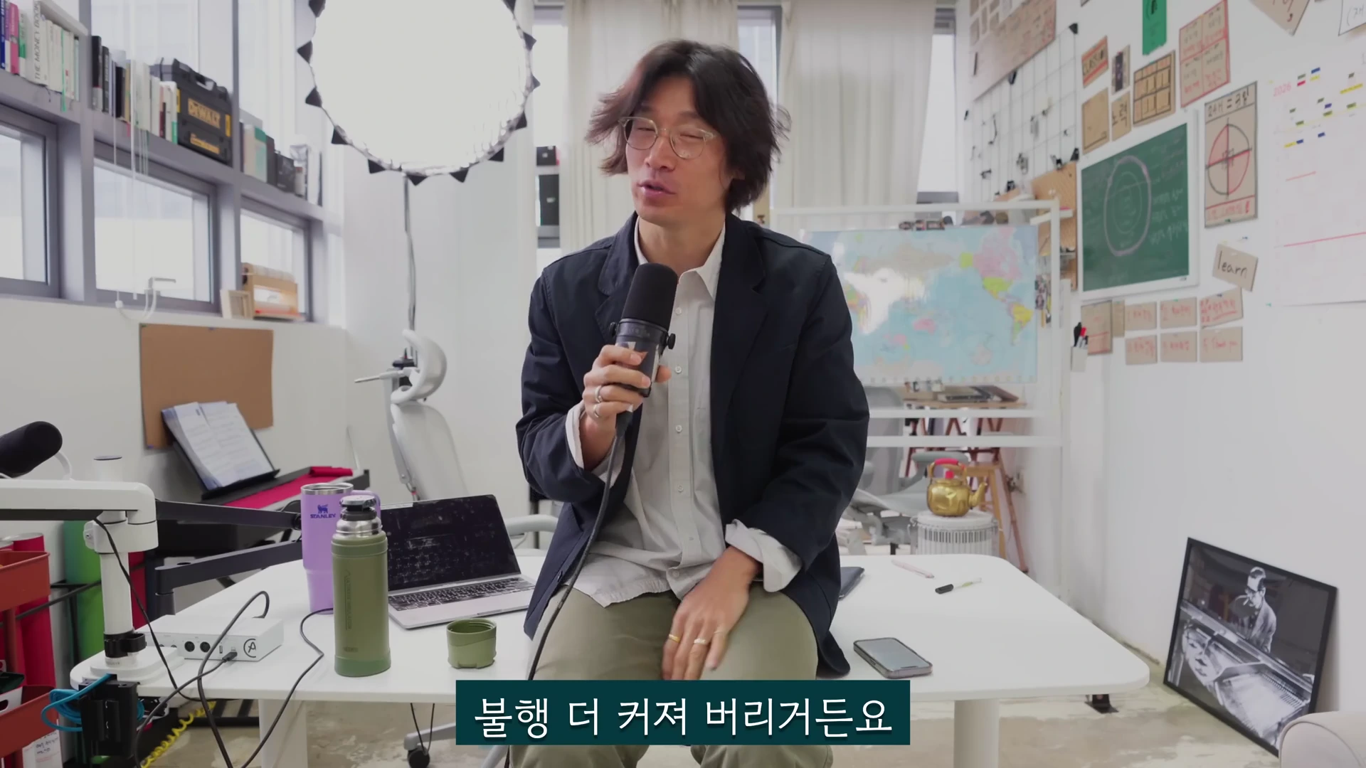 안경을 쓴 남성이 사무실 공간에서 마이크를 들고 앉아 이야기를 하고 있으며, 화면 하단에는 자막이 떠 있다.