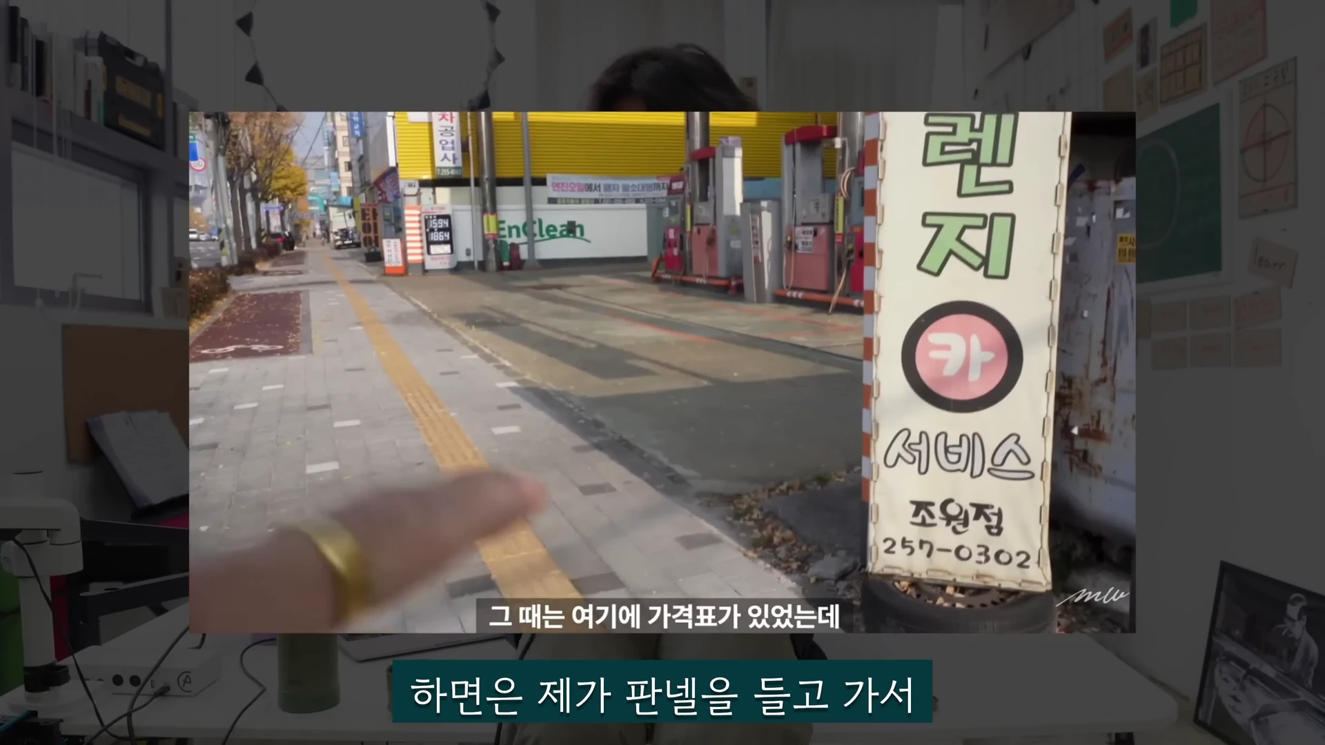 실내 스튜디오에서 화자가 앞에 설치된 화면 속의 주유소 거리와 가격 판넬을 가리키며 설명하고 있다.