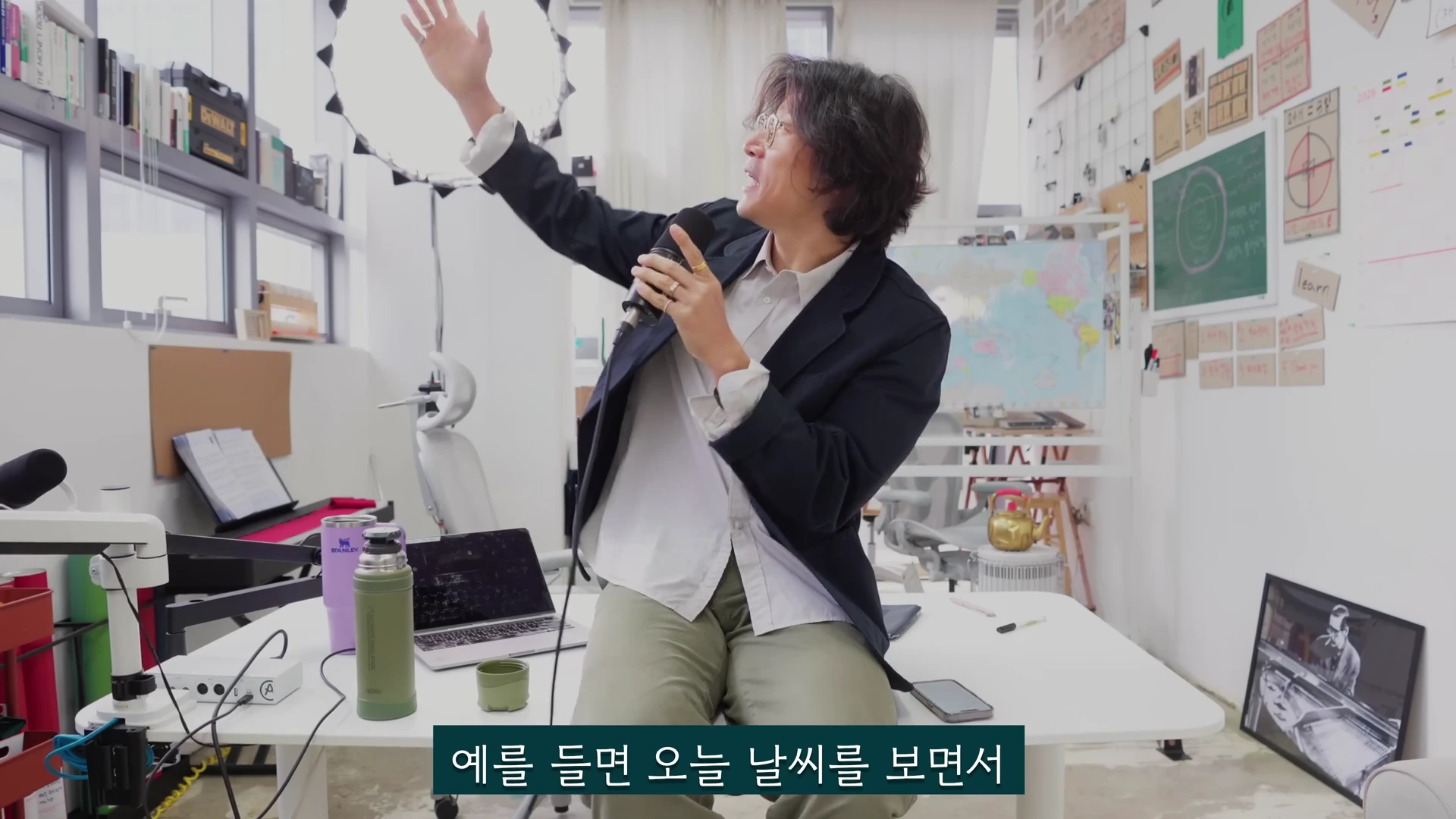 안경을 쓴 남성이 사무실 공간에서 마이크를 들고 한쪽 손을 들어 올리며 이야기하는 모습