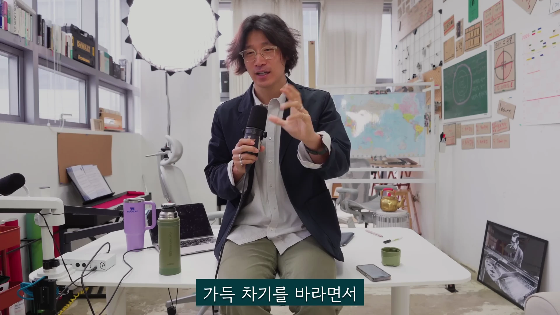 안경을 쓴 남성이 사무실 책상에 앉아 마이크를 들고 손짓하며 이야기를 나누고 있다.