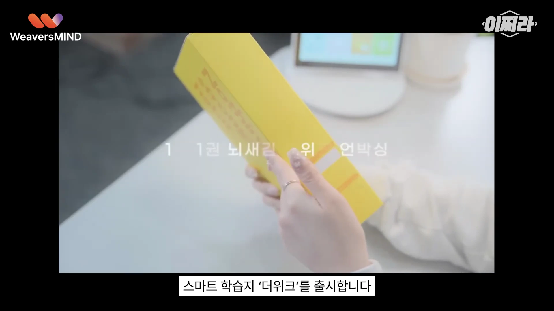 노란색 학습지 패키지를 양손으로 들고 있는 모습이 담긴 영상 캡처 화면이며, 화면 하단에는 스마트 학습지 더위크 출시 자막이 표시되어 있습니다.