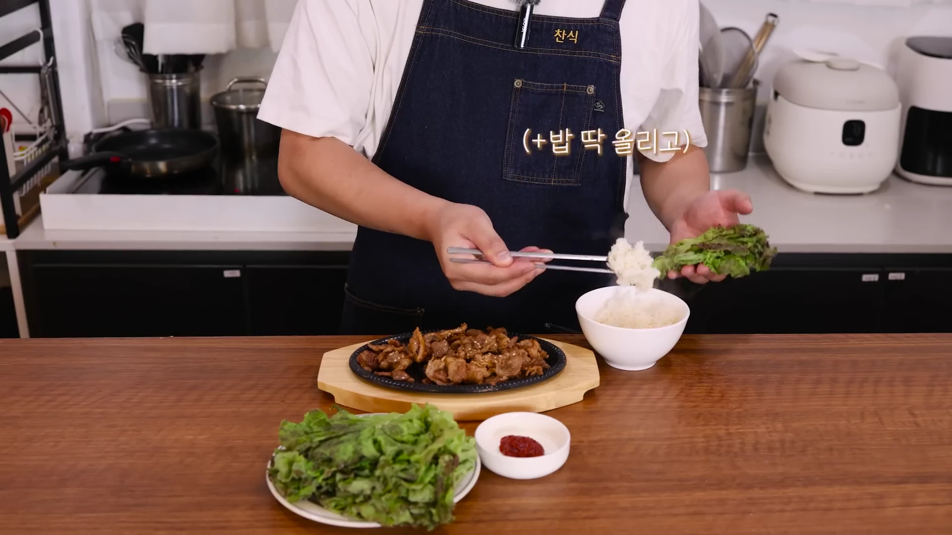 앞치마를 두른 요리사가 상추 위에 밥과 잘 볶아진 돼지고기를 올리며 쌈을 싸는 모습이 담긴 주방 조리대 화면