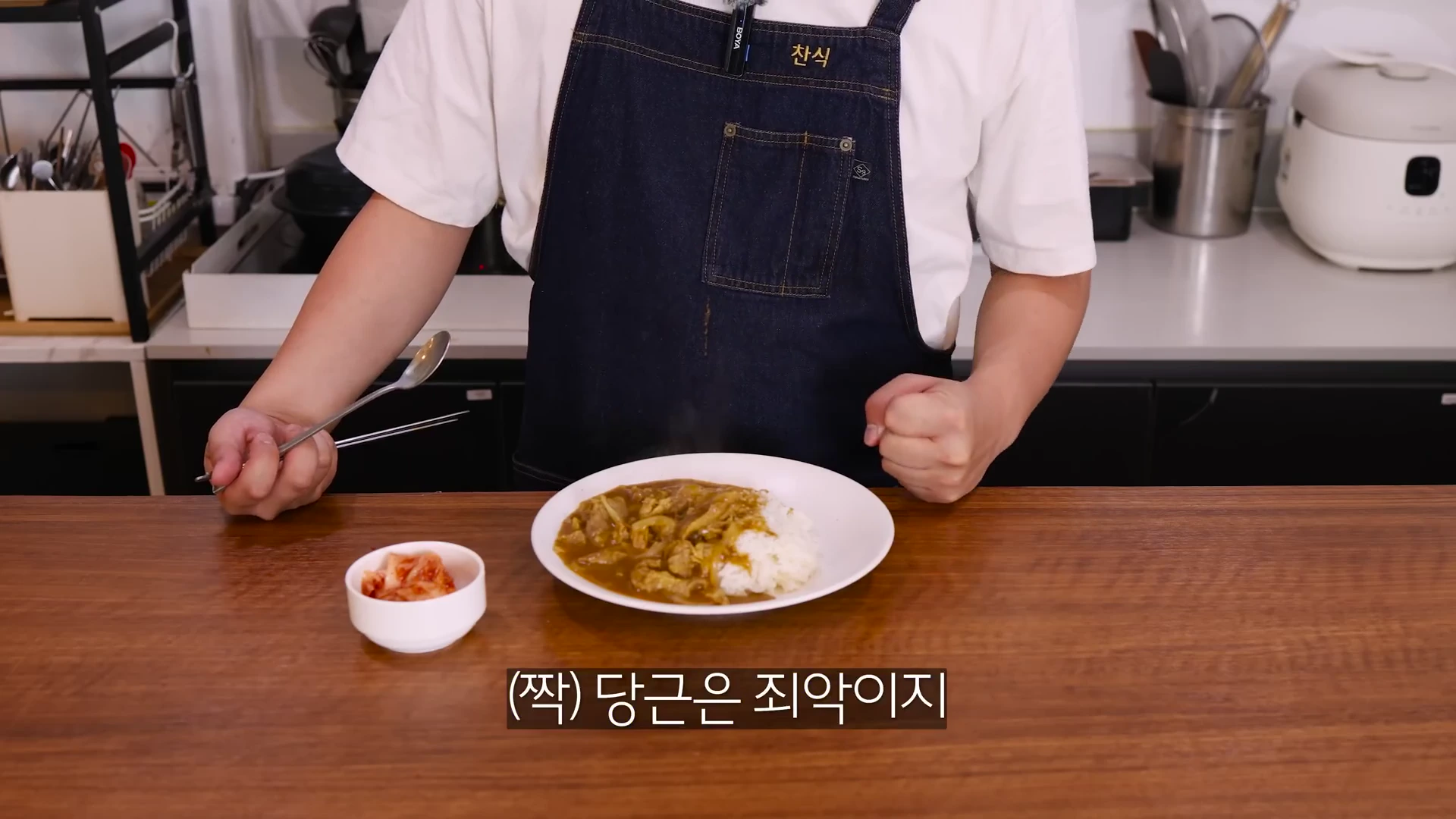 앞치마를 두른 남성이 주방 조리대 앞에서 카레라이스가 담긴 접시를 두고 주먹을 쥐고 있는 모습