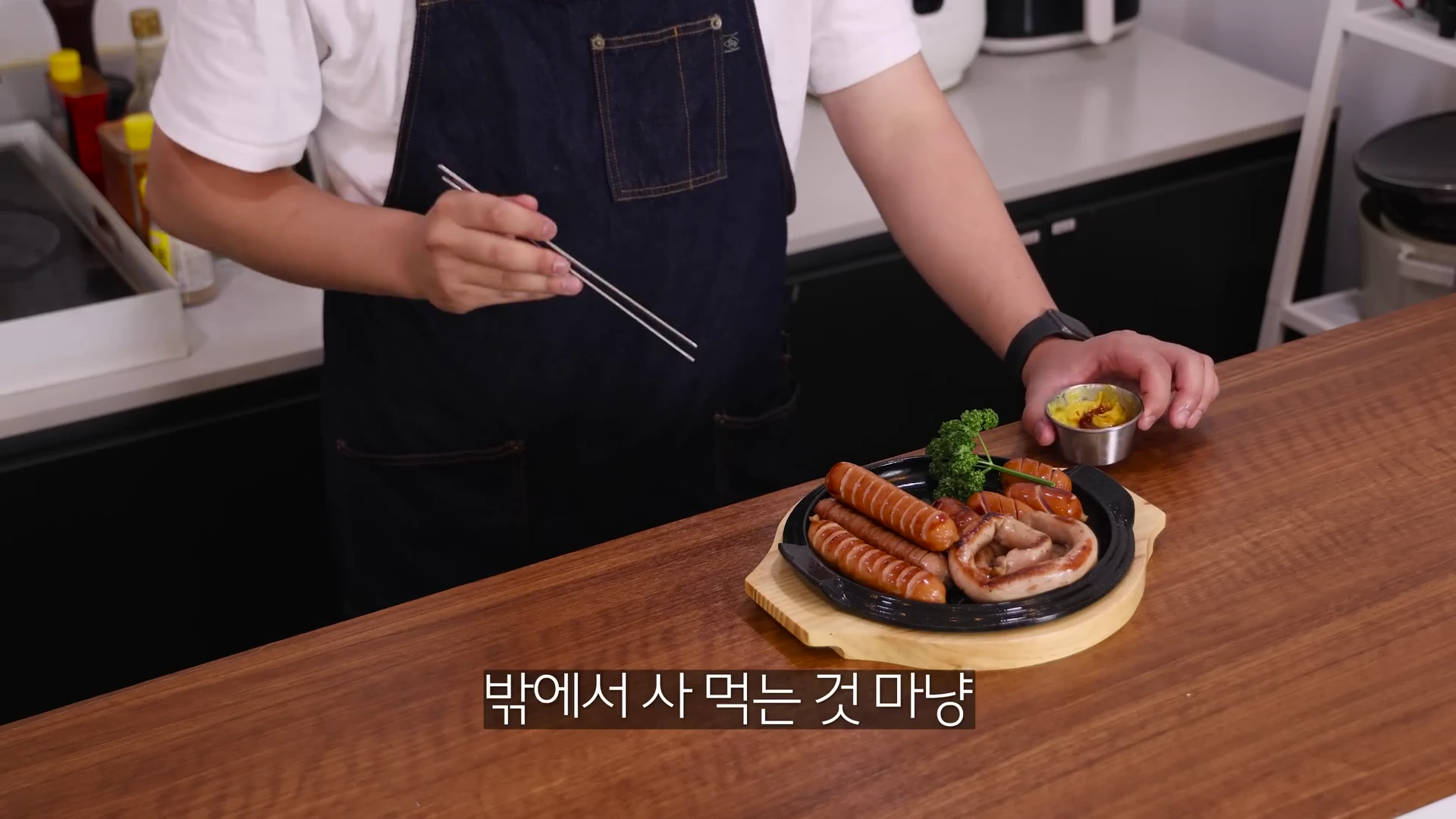 앞치마를 두른 요리사가 칼집을 내어 노릇하게 구운 소시지 접시를 나무 받침대 위에 올려두고 은색 젓가락을 든 모습