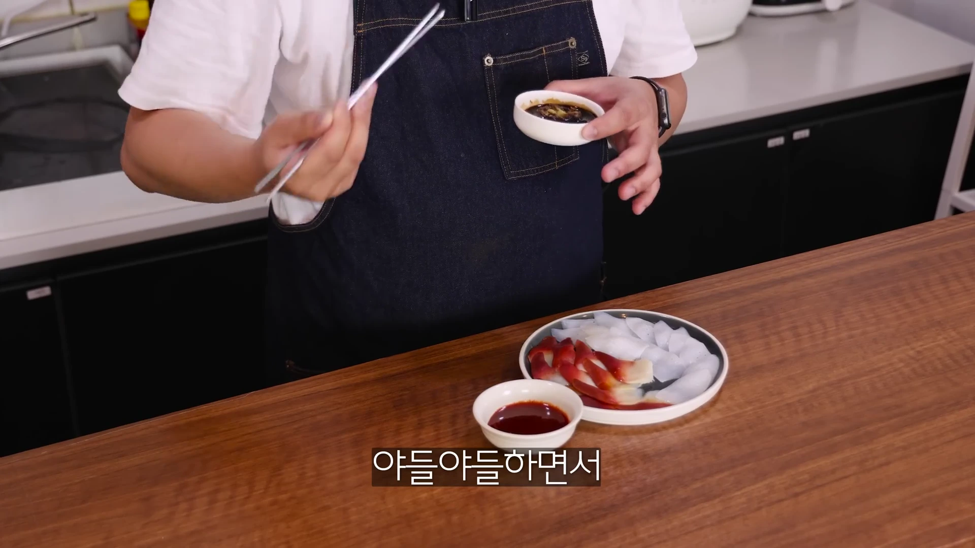 앞치마를 입은 사람이 손에 젓가락과 작은 소스 그릇을 들고, 앞쪽 나무 식탁 위에는 조개 회와 한치 회가 담긴 접시와 초장이 놓여 있다.
