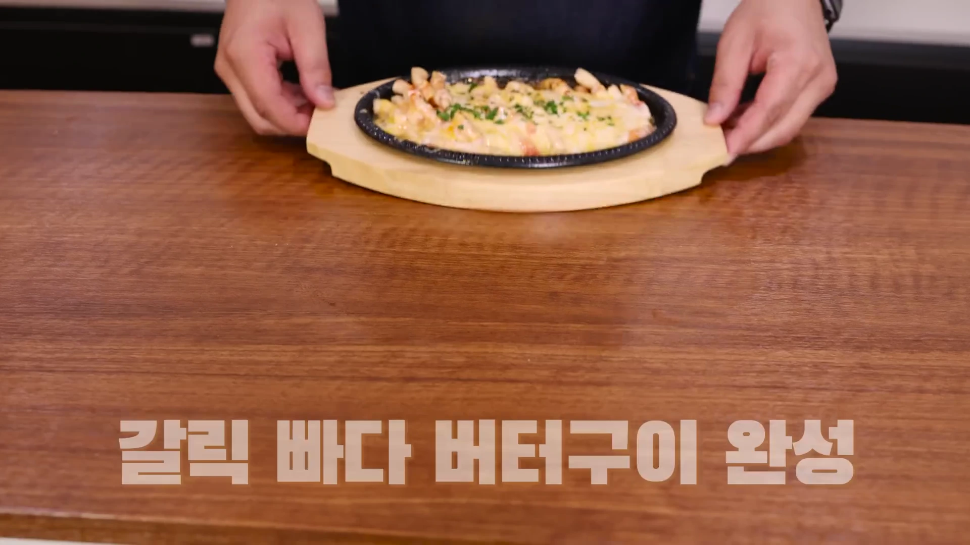 나무 도마 위에 올려진 치즈가 녹아있는 갈릭 버터 대게 다리 요리가 접시에 담겨 있고, 양손으로 이를 들어 올리는 모습이 촬영된 요리 영상의 한 장면입니다.