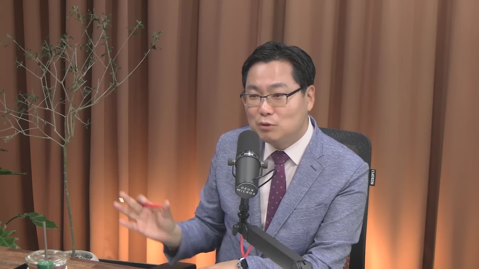 안경을 쓴 남성이 정장 차림으로 스튜디오에서 마이크 앞에 앉아 손짓하며 대화하는 모습