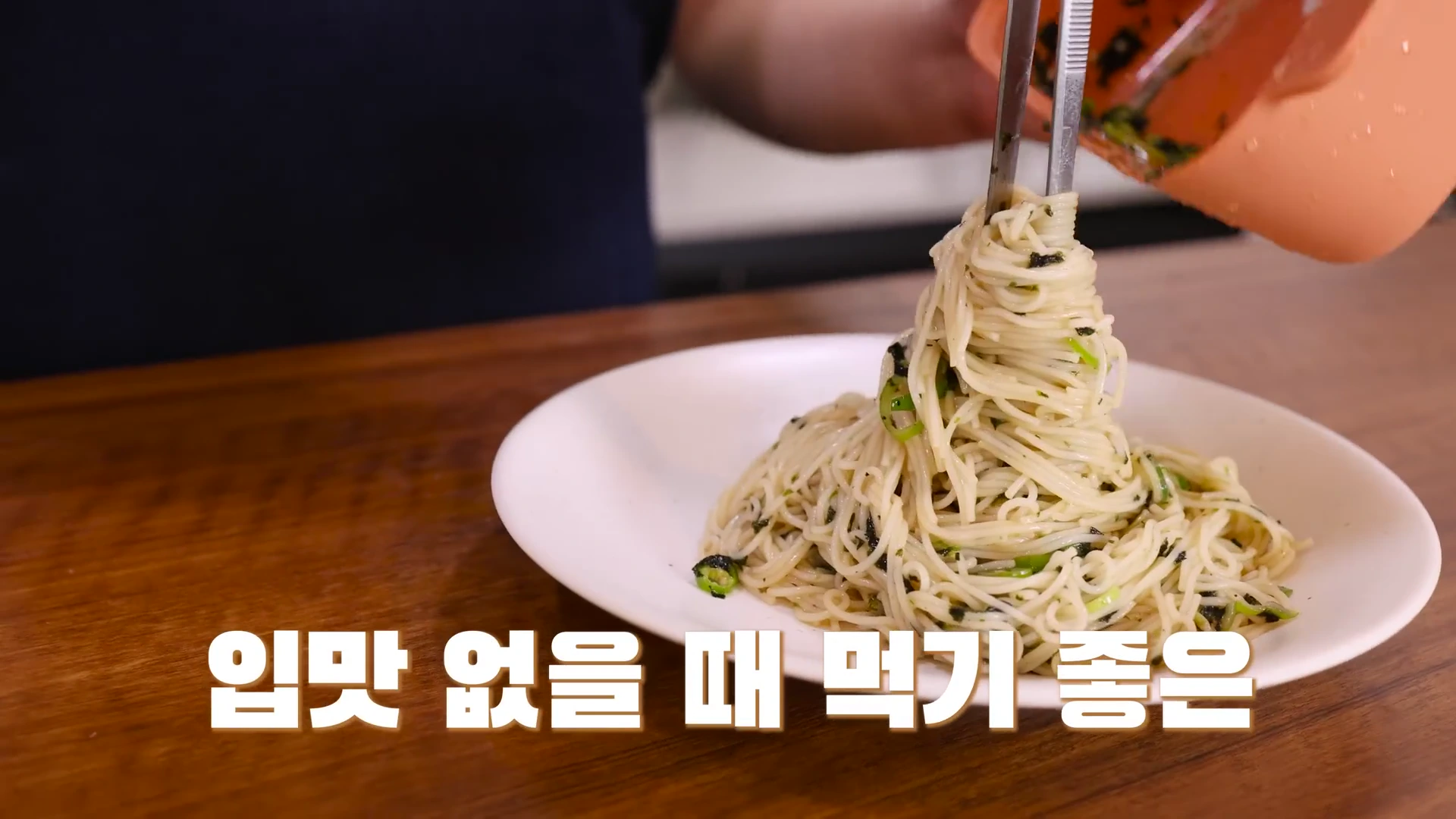 요리용 집게로 비벼진 소면을 들어 올려 흰 접시에 담고 있는 손과 그 아래에 '입맛 없을 때 먹기 좋은'이라는 자막이 놓여 있다.