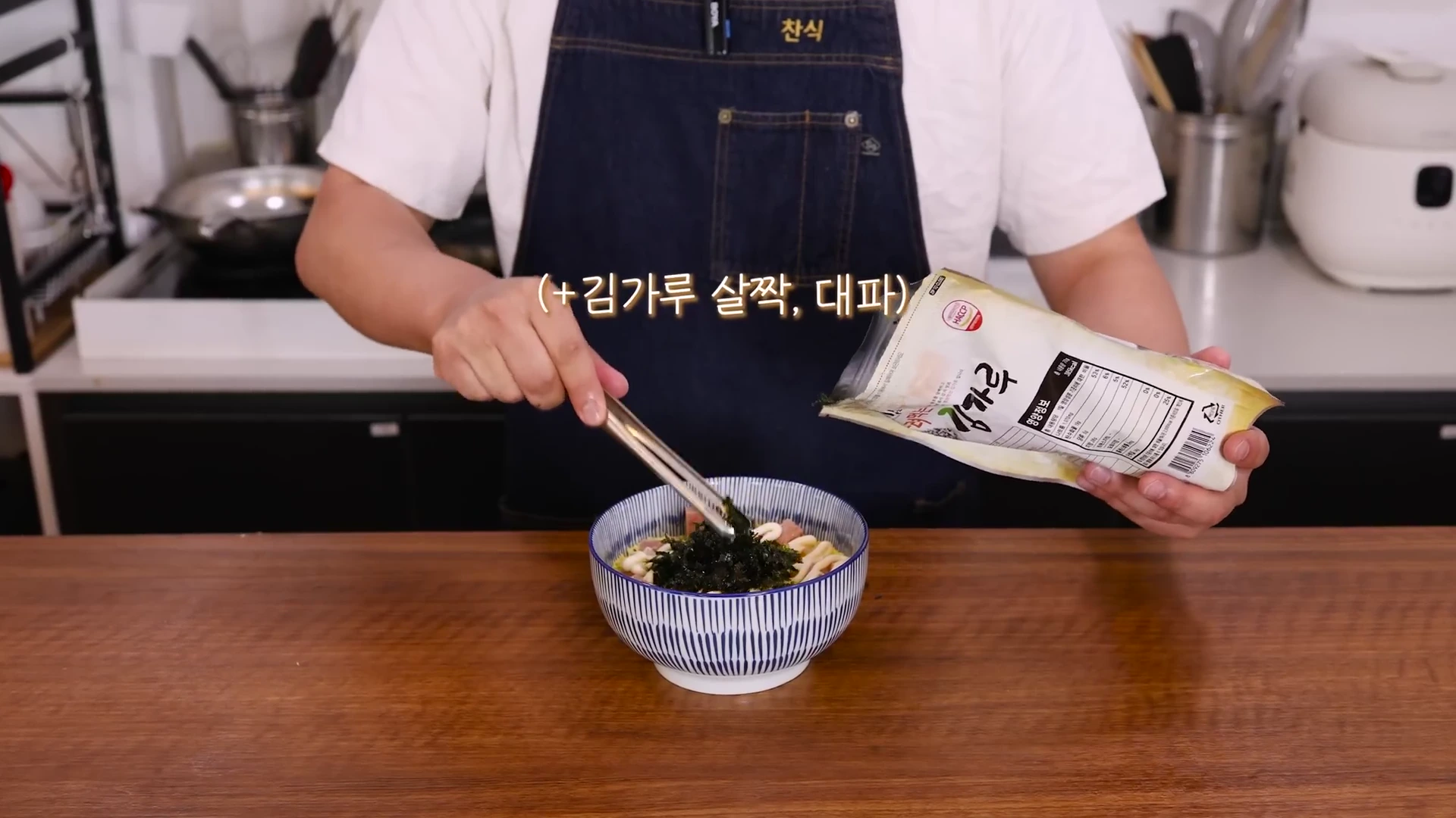 앞치마를 두른 요리사가 주방에서 그릇에 담긴 음식 위에 집게를 이용해 김가루를 뿌리고 있는 모습