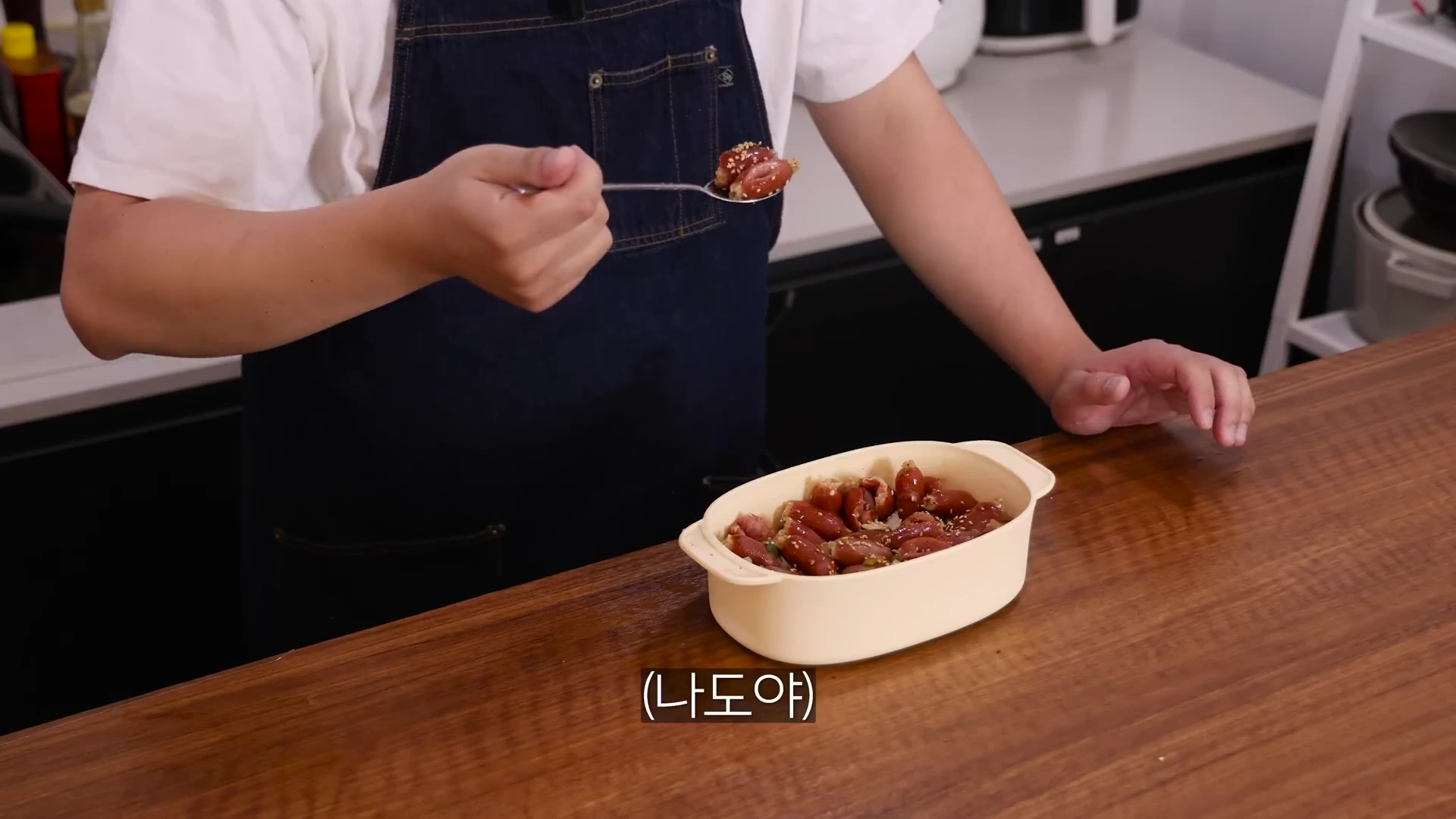 앞치마를 두른 요리사가 숟가락으로 전자레인지 용기에 담긴 소시지 야채 볶음을 들어 올리고 있는 모습이다.