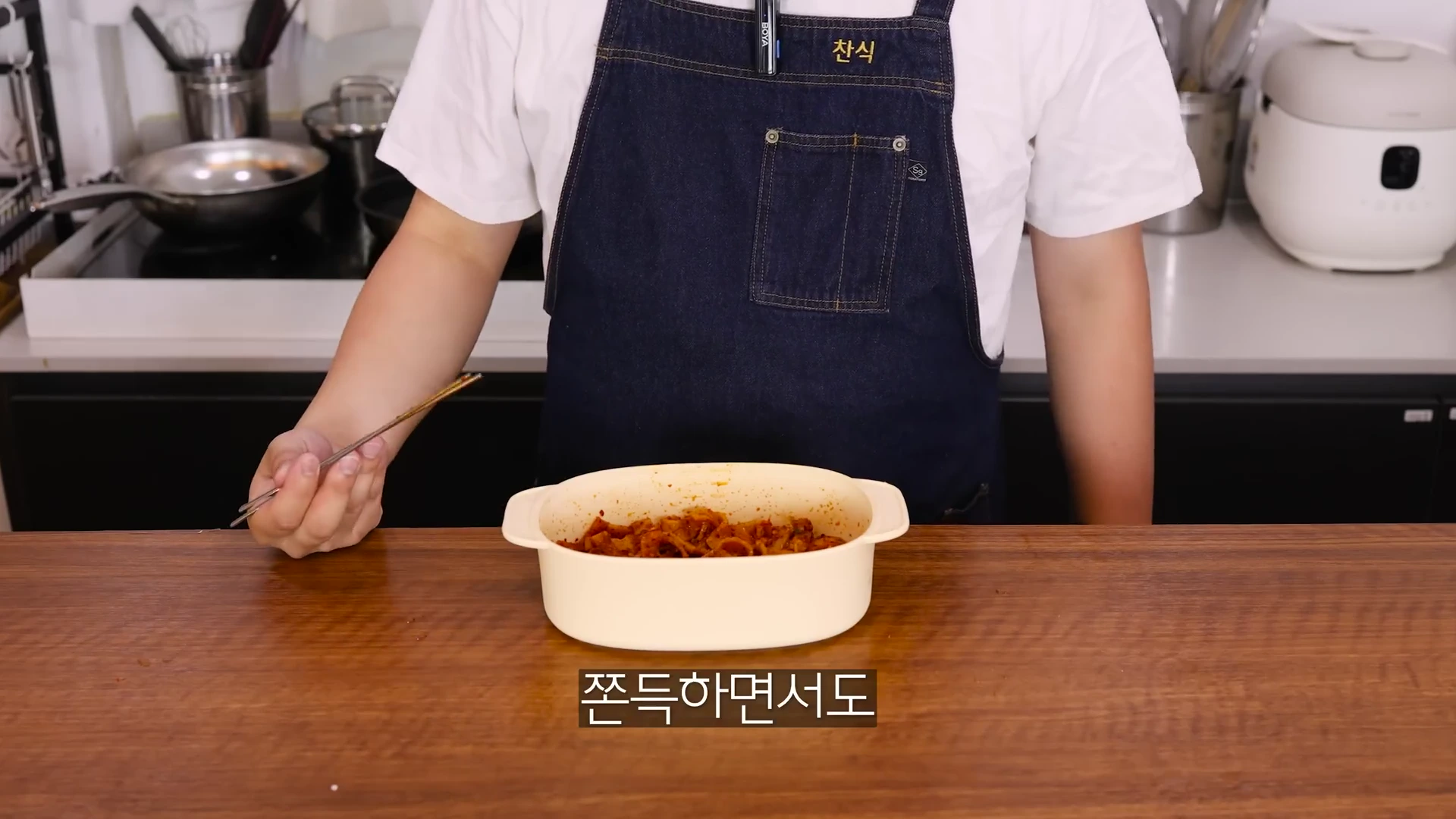 흰 티셔츠에 앞치마를 두른 요리사가 전자레인지 전용 용기에 담긴 완성된 어묵 요리를 앞에 두고 젓가락을 들고 서 있다.