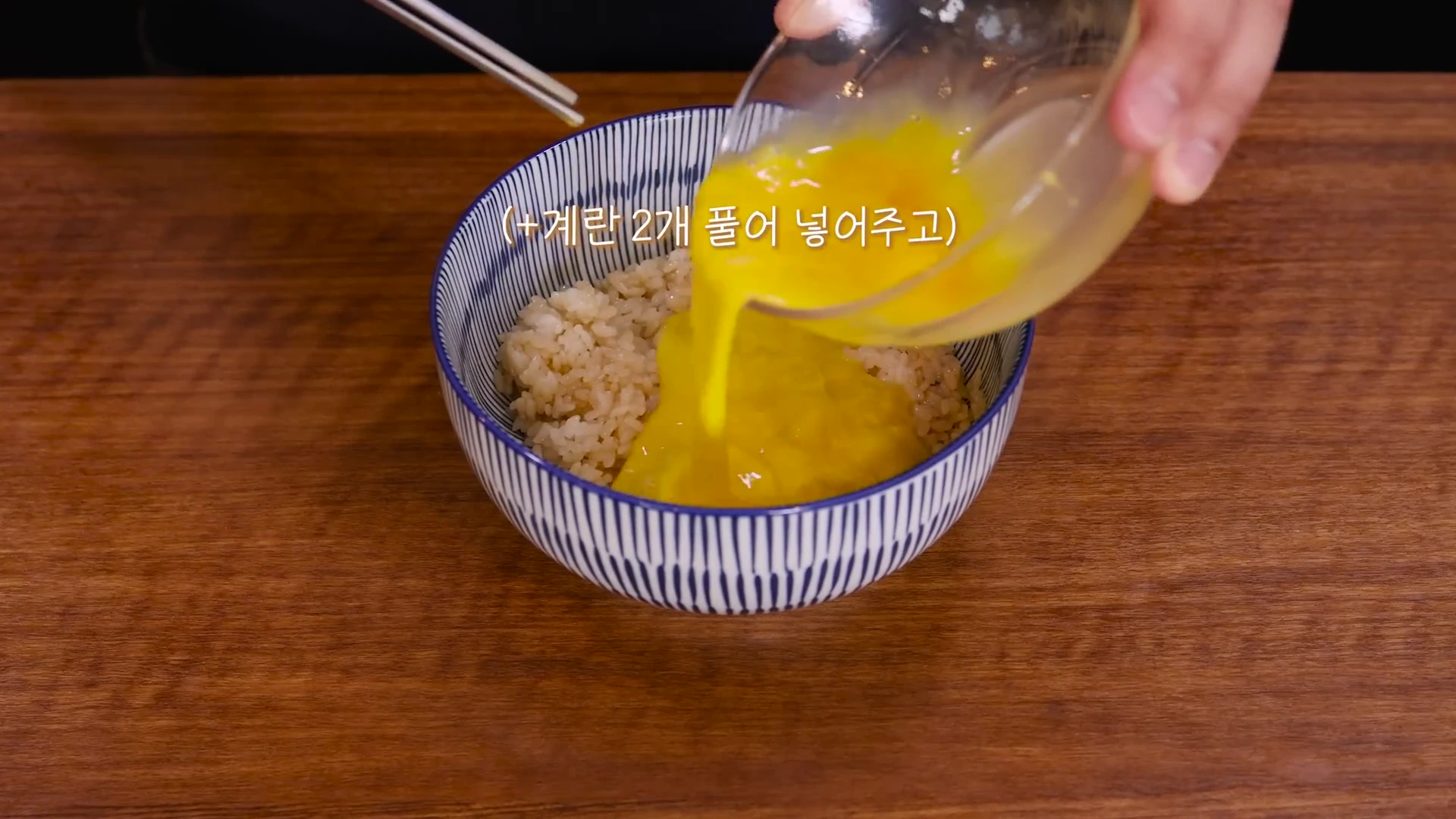 나무 식탁 위에 놓인 파란색 줄무늬 그릇에 담긴 밥 위로 투명한 유리그릇에 든 푼 달걀물을 붓는 모습이며 화면 중앙에는 '+계란 2개 풀어 넣어주고'라는 한국어 자막이 적혀 있다.