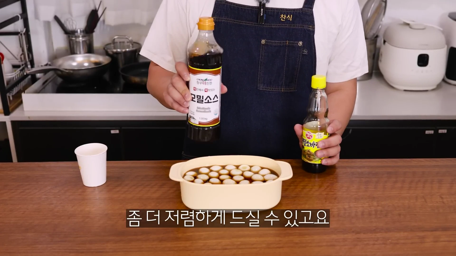 앞치마를 입은 요리사가 양손에 메밀 소스 병을 들고 전자레인지 용기에 담긴 메추리알 장조림 재료를 보여주는 모습