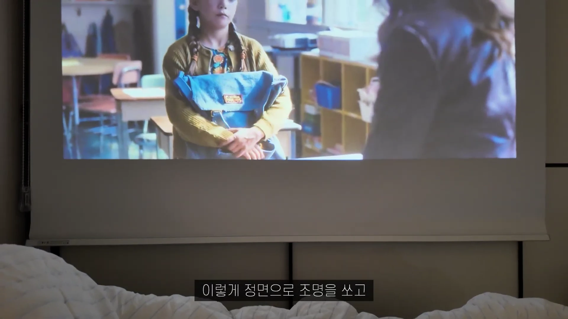 빔프로젝터로 투사된 영화 화면이 스크린에 선명하게 비춰지고 있으며 하단에는 자막이 포함되어 있습니다.
