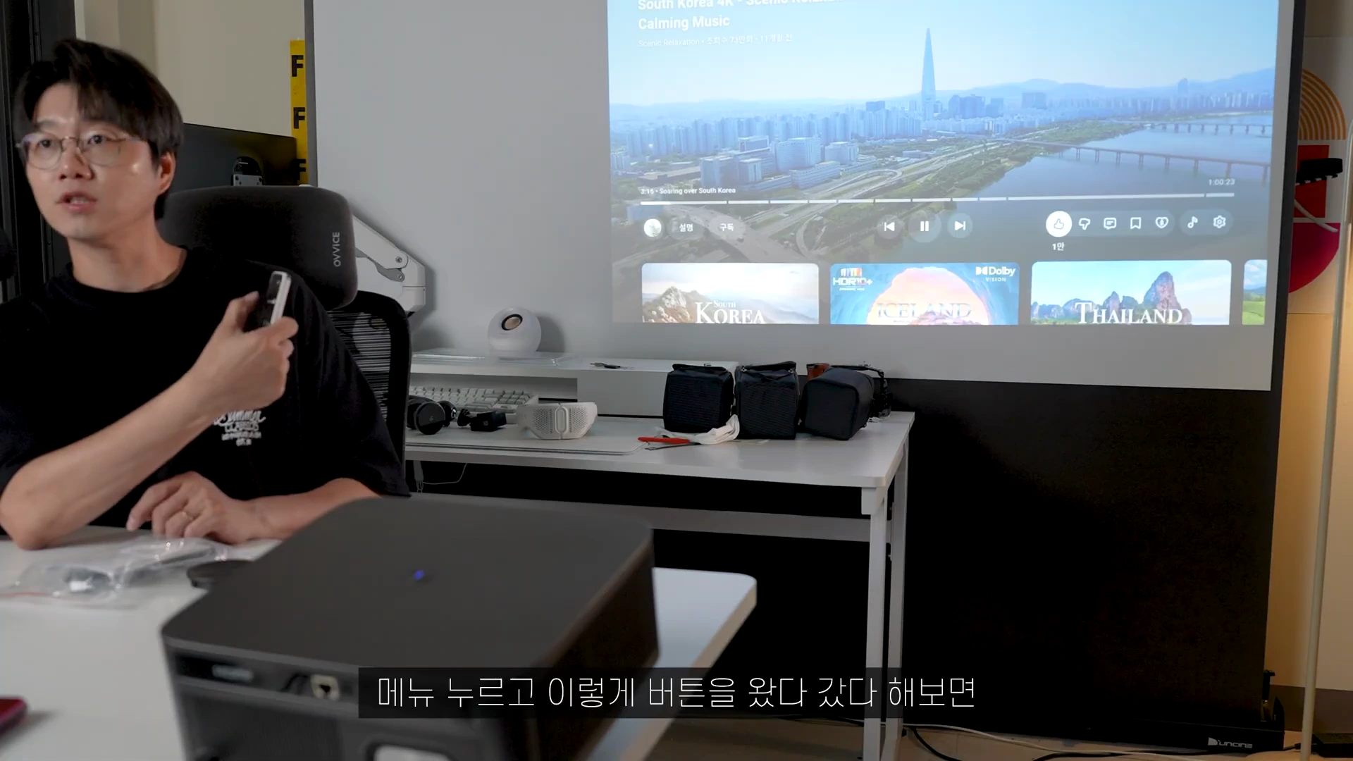 안경을 쓴 남성이 리모컨을 들고 빔프로젝터로 투사된 화면을 보며 설명하고 있는 모습.