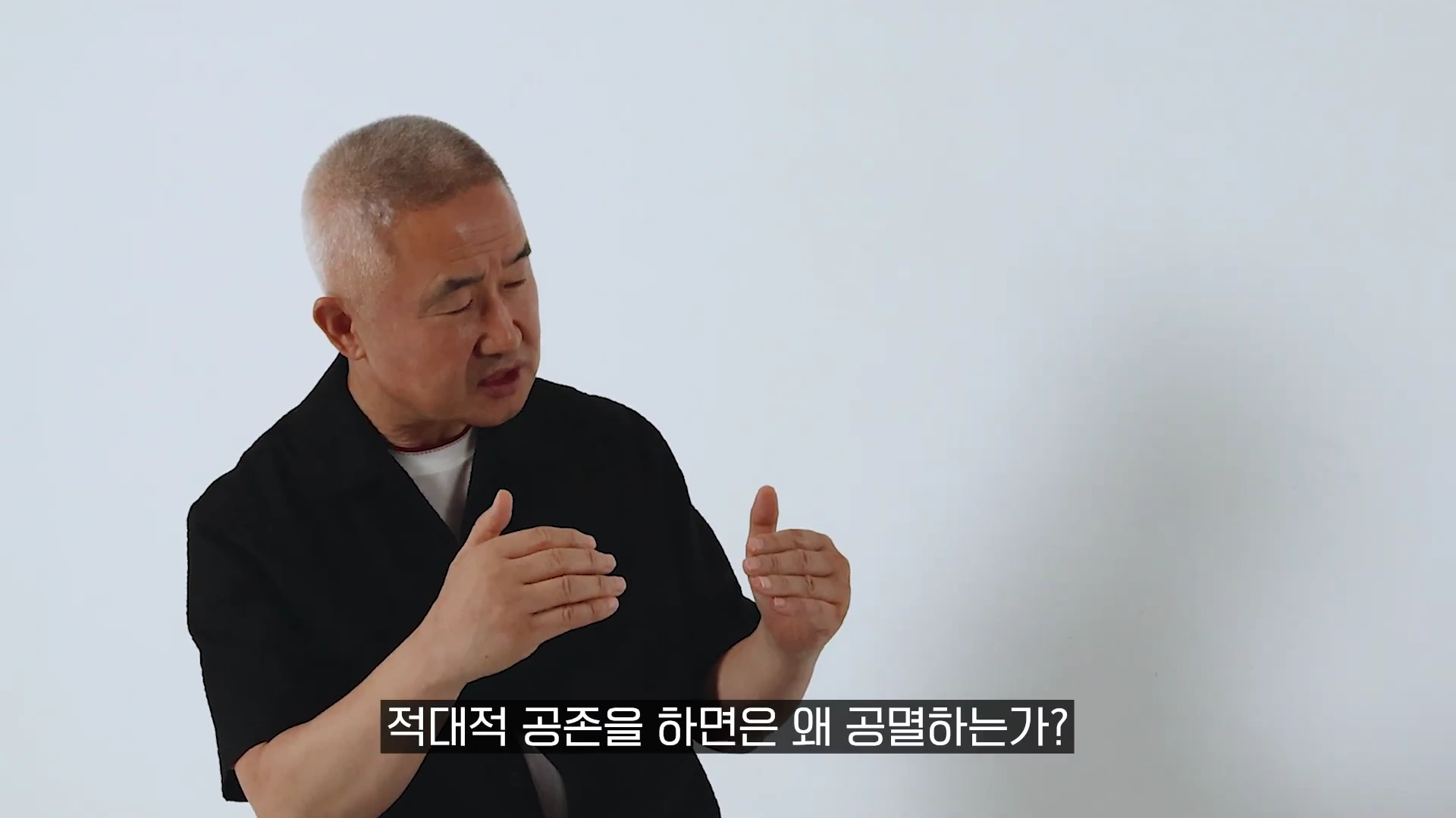 흰 배경 앞에 앉아 검은색 상의를 입은 중년 남성이 손동작을 하며 이야기를 하고 있으며 화면 하단에는 적대적 공존을 하면은 왜 공멸하는가?라는 자막이 표시되어 있다.