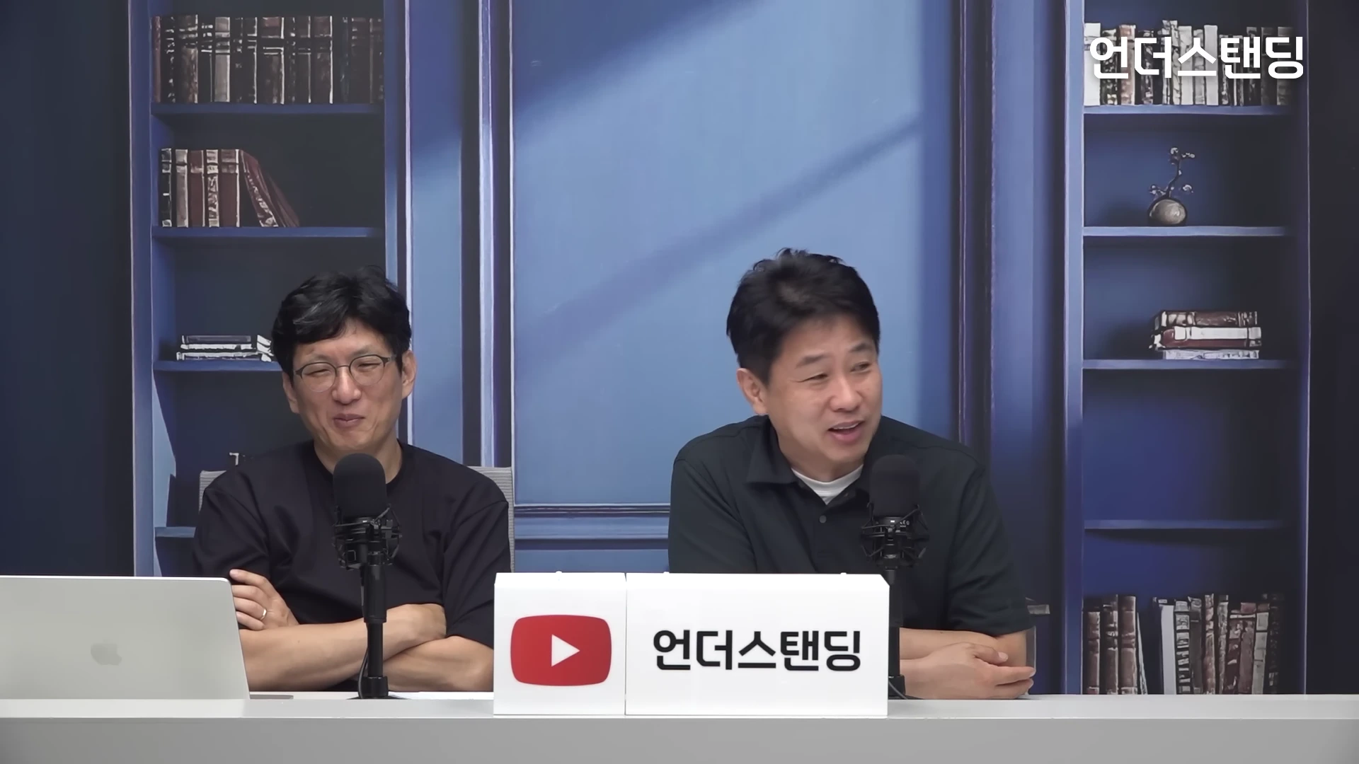 두 명의 남성이 책장 배경의 스튜디오에서 마이크를 앞에 두고 앉아 대화를 나누고 있다.