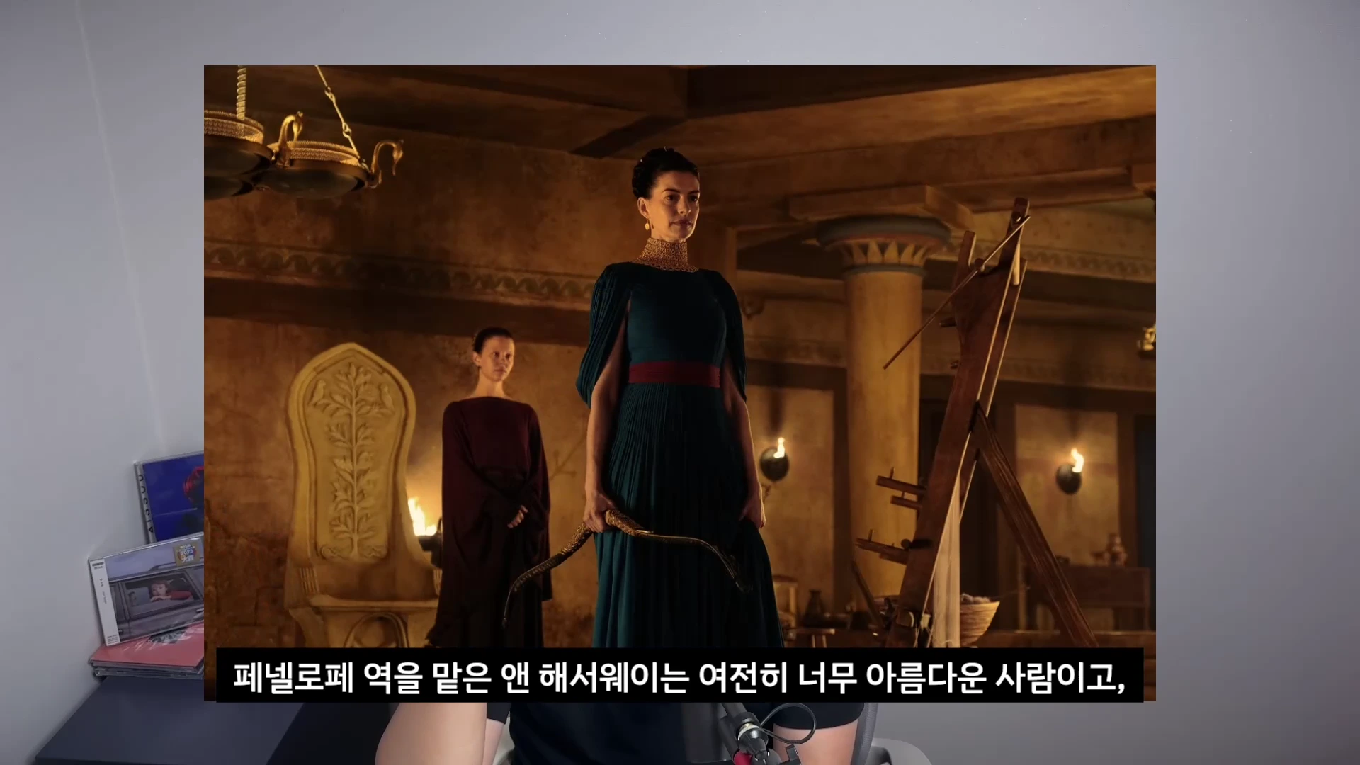 영화 속 어두운 고대 궁전 배경에서 짙은 청록색 드레스를 입고 활을 든 여성이 정면을 응시하고 있으며 뒤편에는 다른 여성이 서 있는 모습