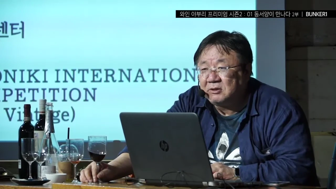 안경을 쓴 중년 남성이 노트북 앞에서 와인 잔을 들고 강연하는 모습. 앞에는 와인 병과 잔이 놓여 있고 뒤쪽 스크린에는 영어 텍스트가 적힌 슬라이드가 비치고 있음.