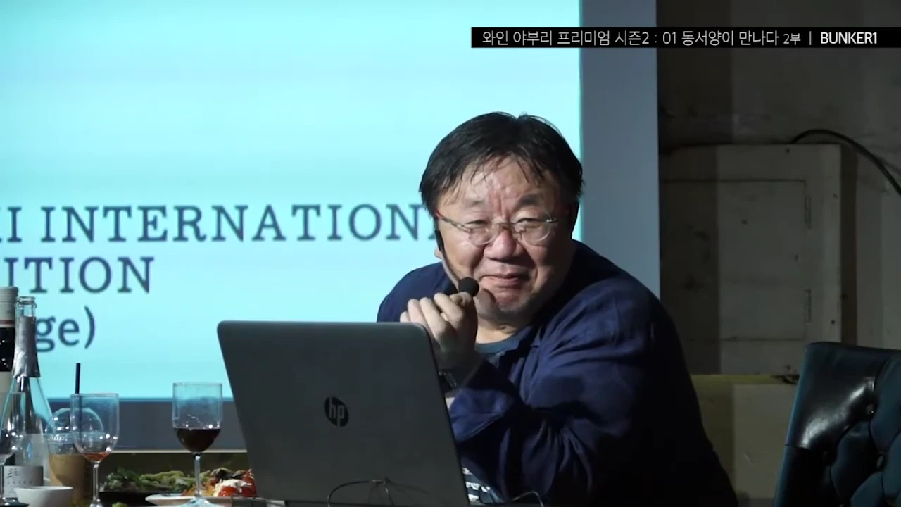 A man with glasses smiling and talking during a presentation in front of a projection screen with text, sitting at a desk with wine glasses and a laptop.