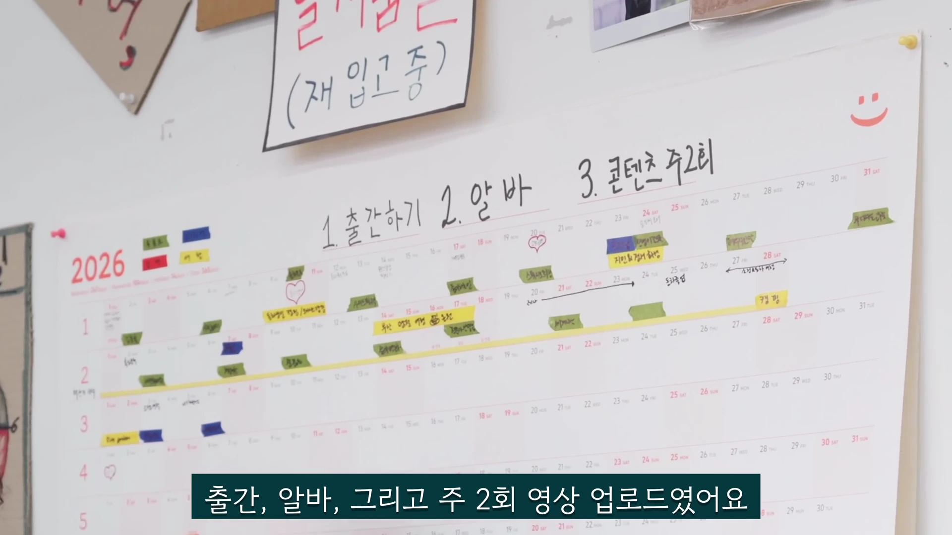 2026년 연간 계획이 적힌 대형 달력 벽보에 1번 출간하기, 2번 알바, 3번 콘텐츠 주 2회 업로드라는 손글씨 목표가 적혀 있다.
