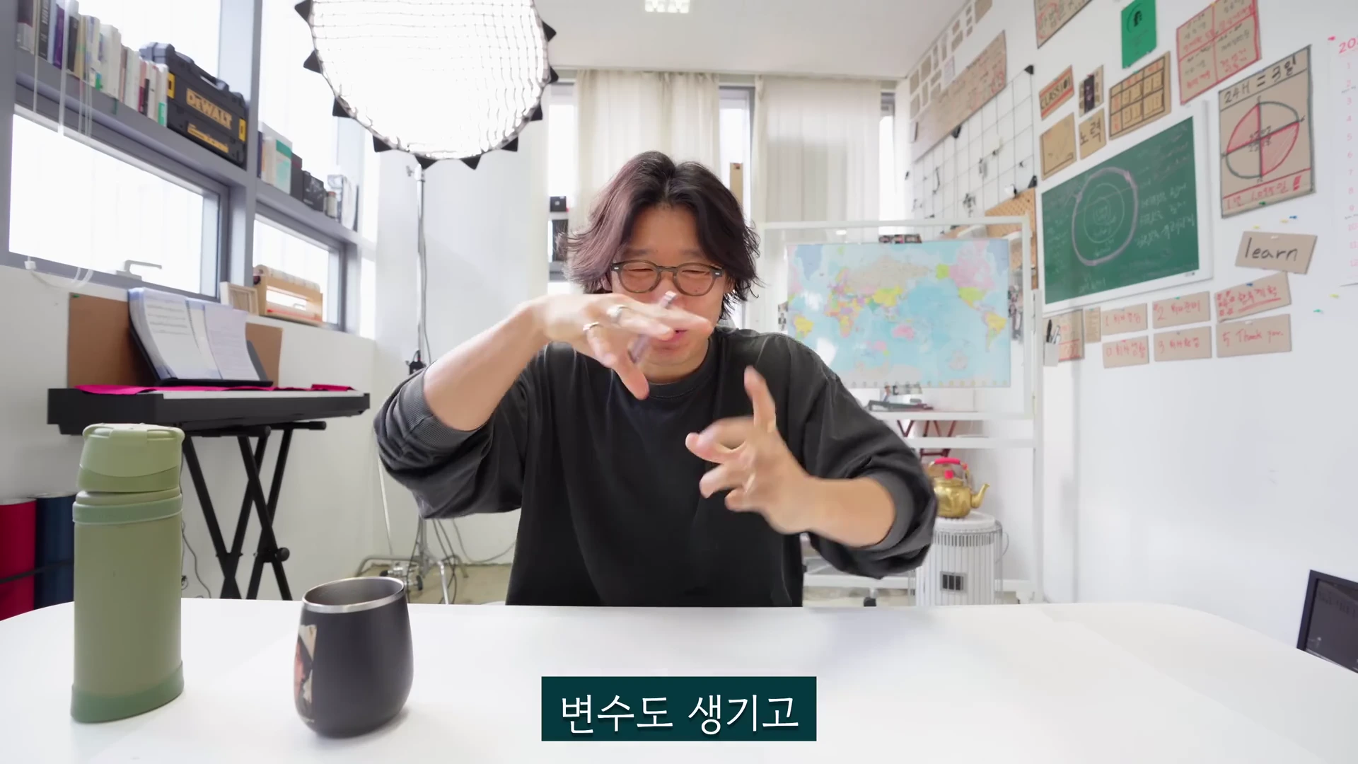 안경을 쓴 남성이 책상에 앉아 양손으로 무언가를 설명하는 듯한 동작을 취하고 있으며, 배경의 벽면에는 세계 지도와 목표가 적힌 종이들이 붙어 있습니다.