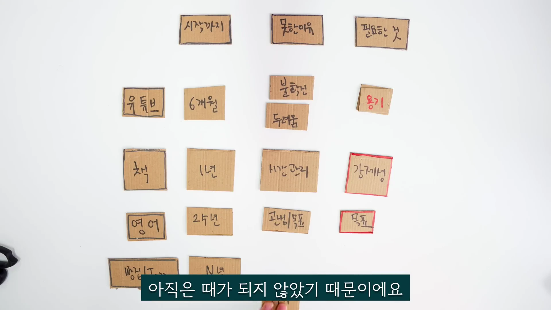흰색 배경 위에 '시작하지 못한 이유', '필요한 것'과 같은 문구가 적힌 골판지 조각들이 행과 열에 맞춰 배열되어 있고, 하단에는 '아직은 때가 되지 않았기 때문이에요'라는 자막이 입혀져 있다.