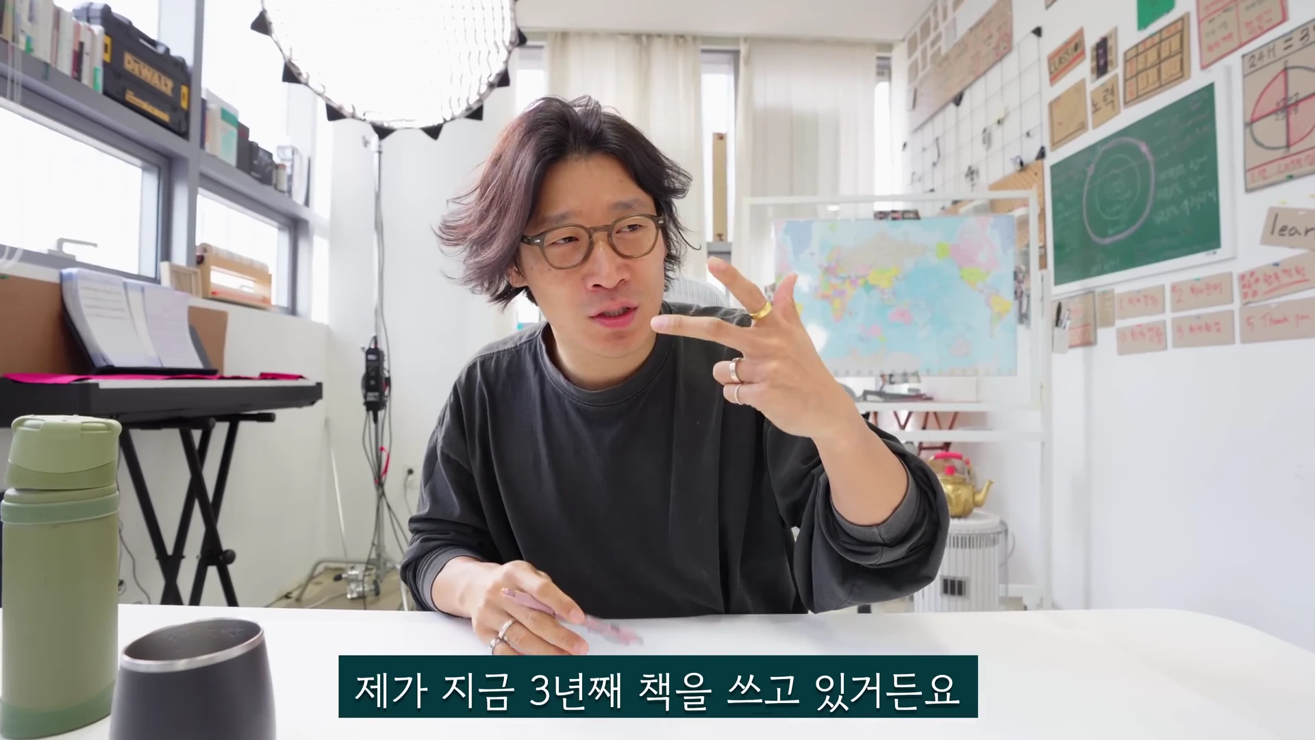 밝은 작업실 배경의 책상 앞에 앉아 안경을 쓴 남성이 손가락으로 숫자를 표시하며 이야기하고 있는 모습